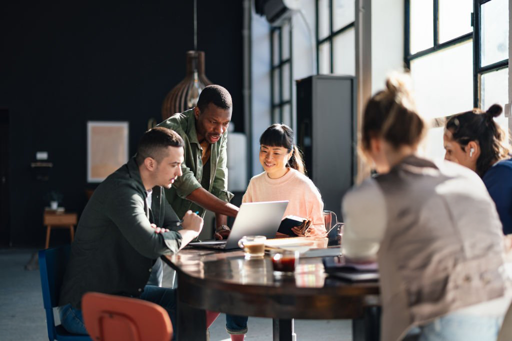 A diverse group of five young adults working together in a modern office space with large windows, gathered around a table with a laptop and notebooks, engaged in discussion.