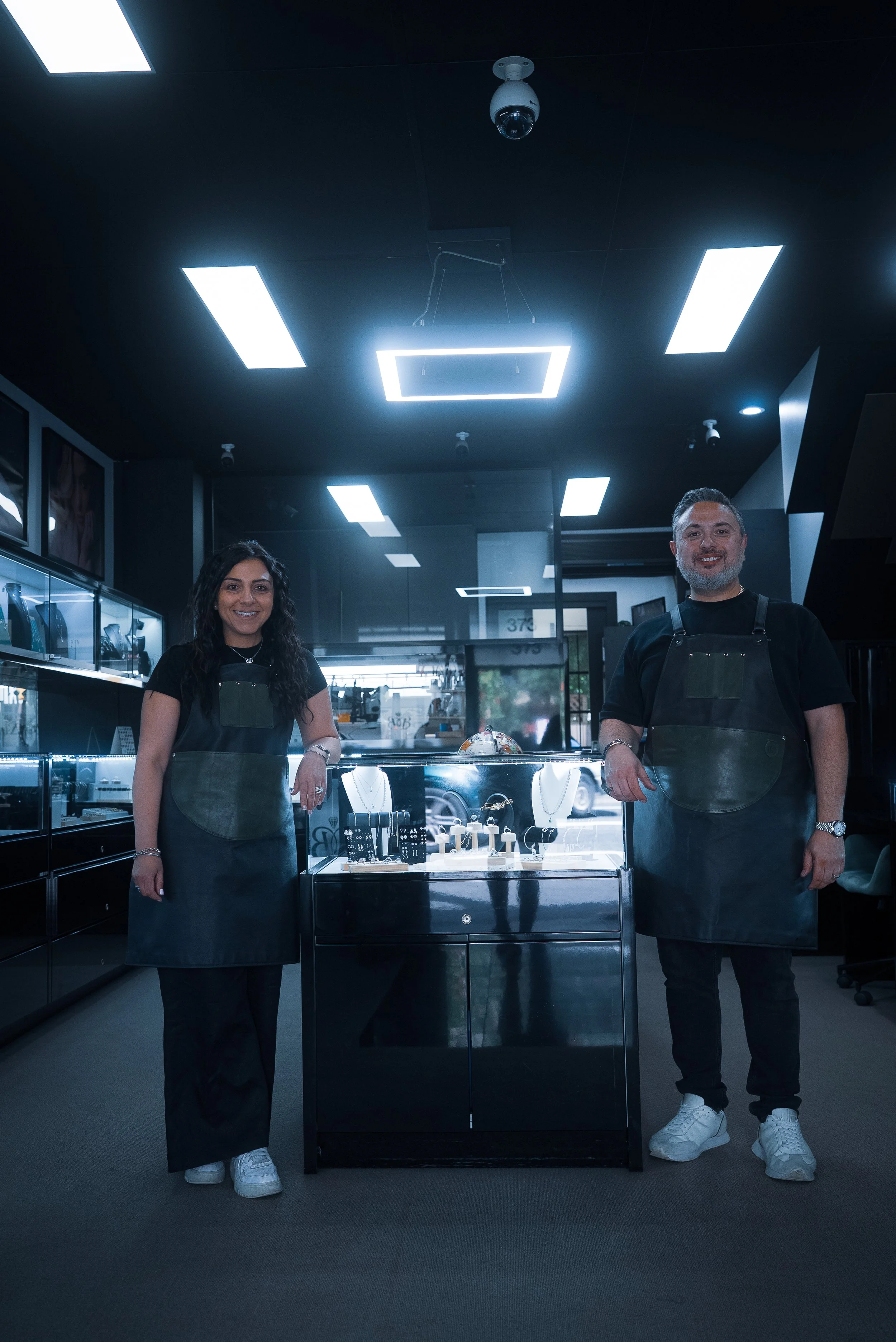 Two jewelry store employees, a woman and a man, standing behind a display case with jewelry, in a modern, dark-colored interior.