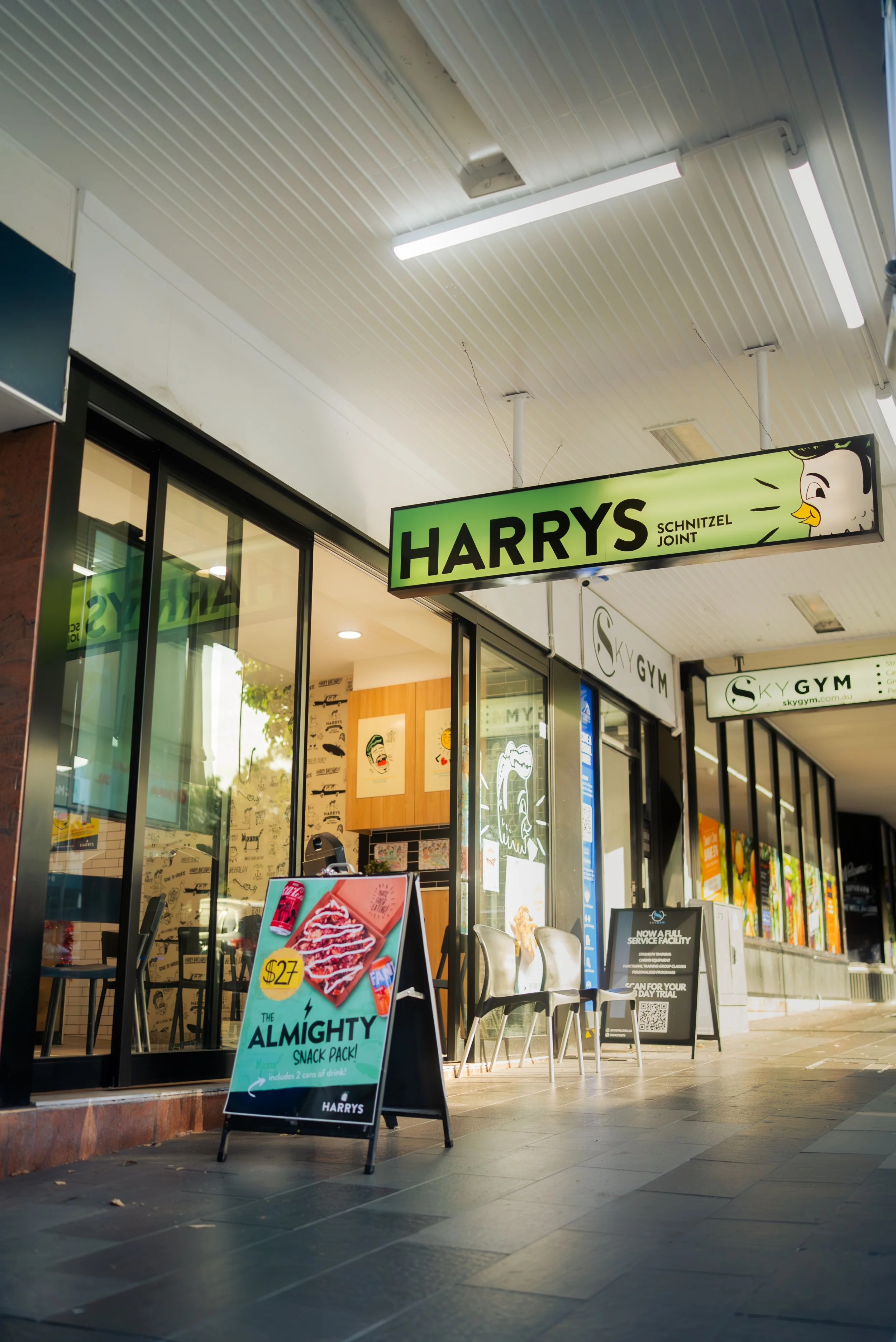 Street view of Harry's sandwich shop with green sign and logo, displaying a cartoon chicken, an advertisement board showing a snack pack for $27, and chairs outside.