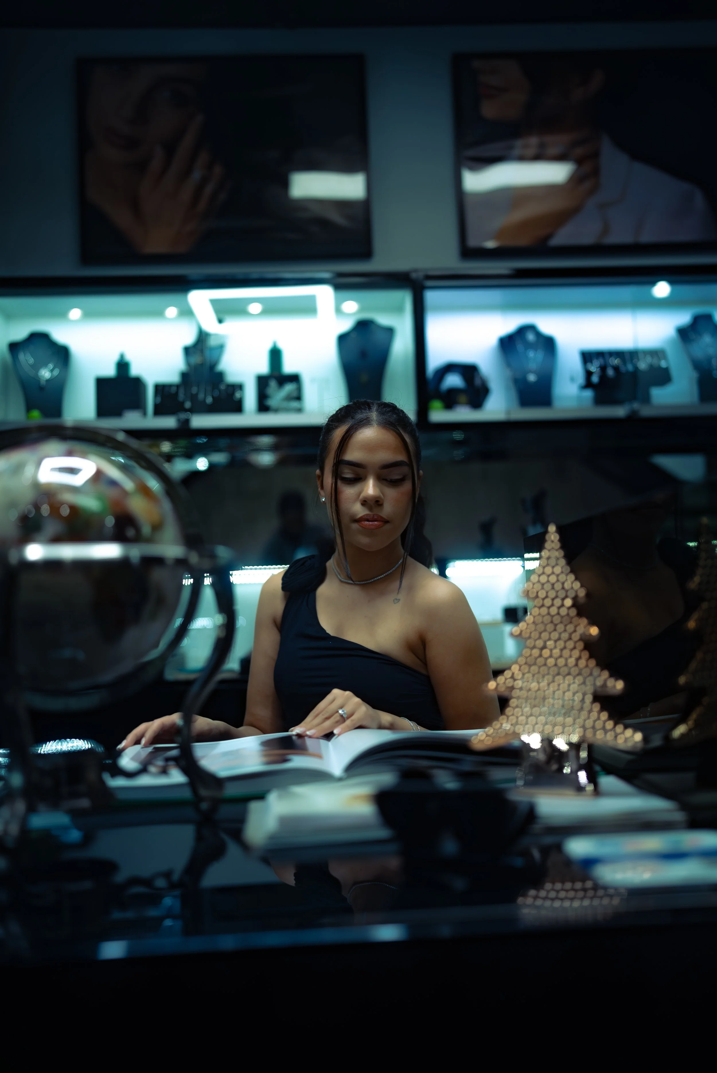 Young woman with dark hair studying a book in a jewelry store, with jewelry display cases and posters of women on the wall behind her.