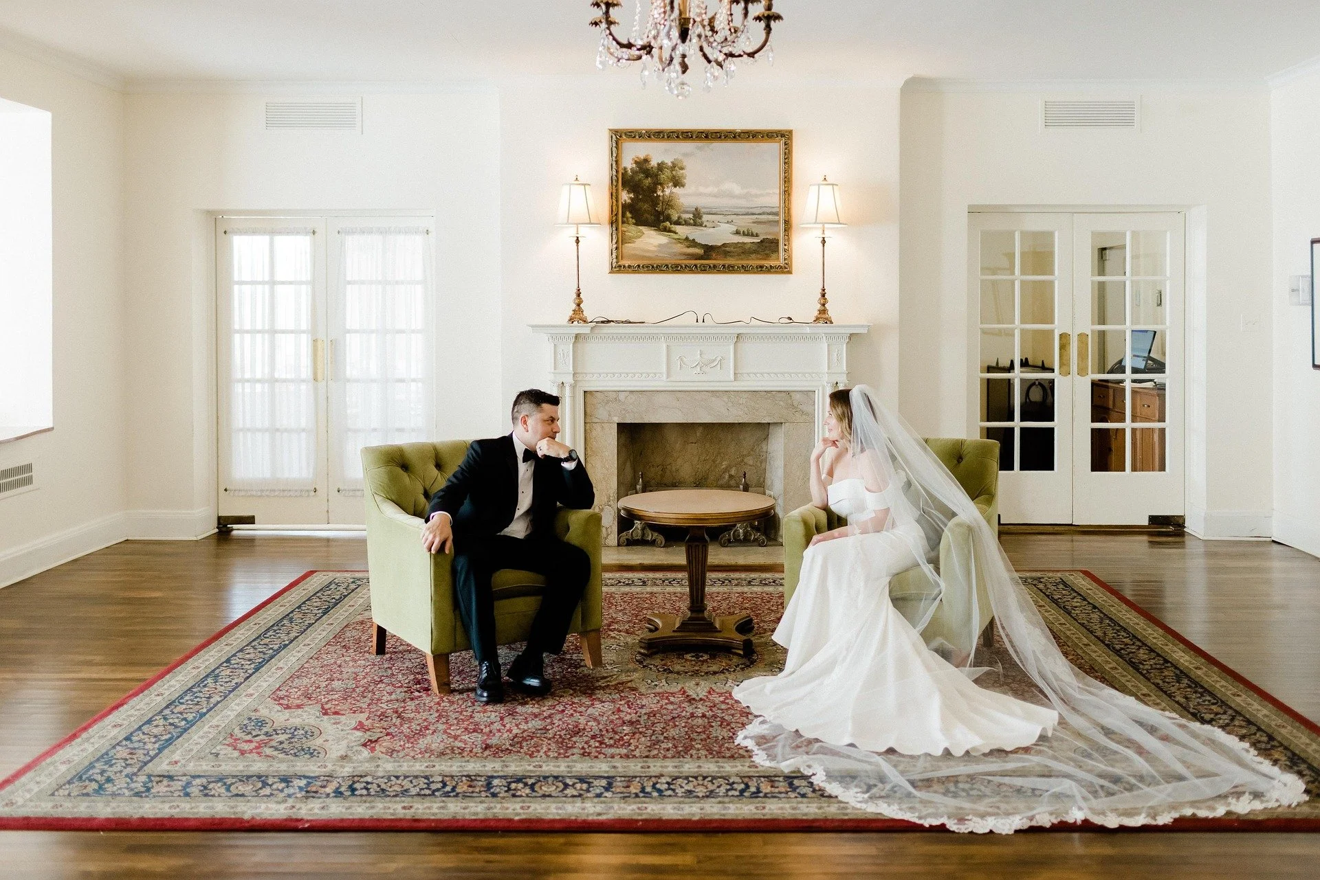 A bride and groom sitting in a living room, facing each other. The bride is wearing a white wedding dress with a veil, and the groom is in a black tuxedo. The room has a fireplace, a chandelier, and a large area rug.