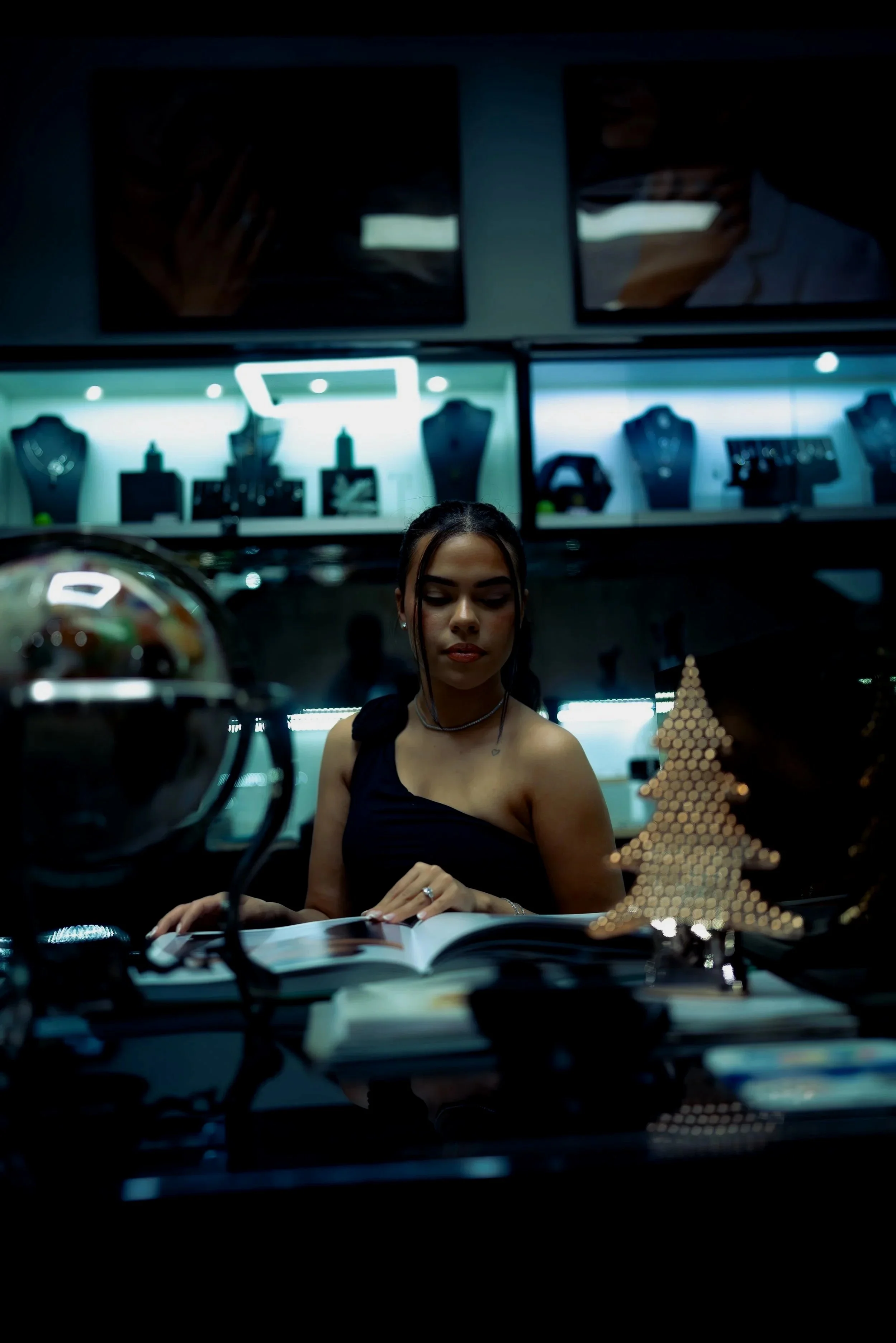 A young woman in a black dress sitting at a desk, reading a book, in a jewelry store with display cases of jewelry and a small decorative Christmas tree on the desk.