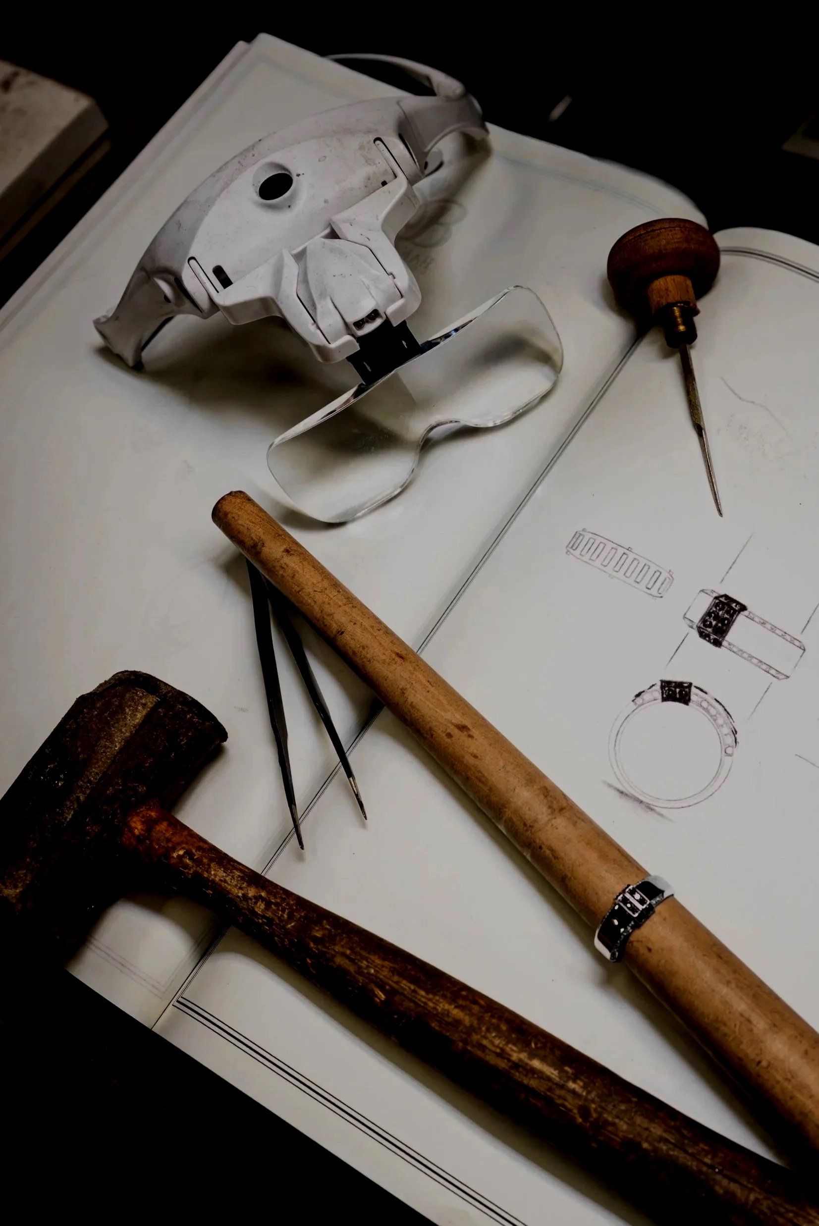Tools and a technical drawing on a table, including a white robotic arm, a screwdriver with a wooden handle, a hammer, a pair of tweezers, and a wrench, along with a sketch of a watch.