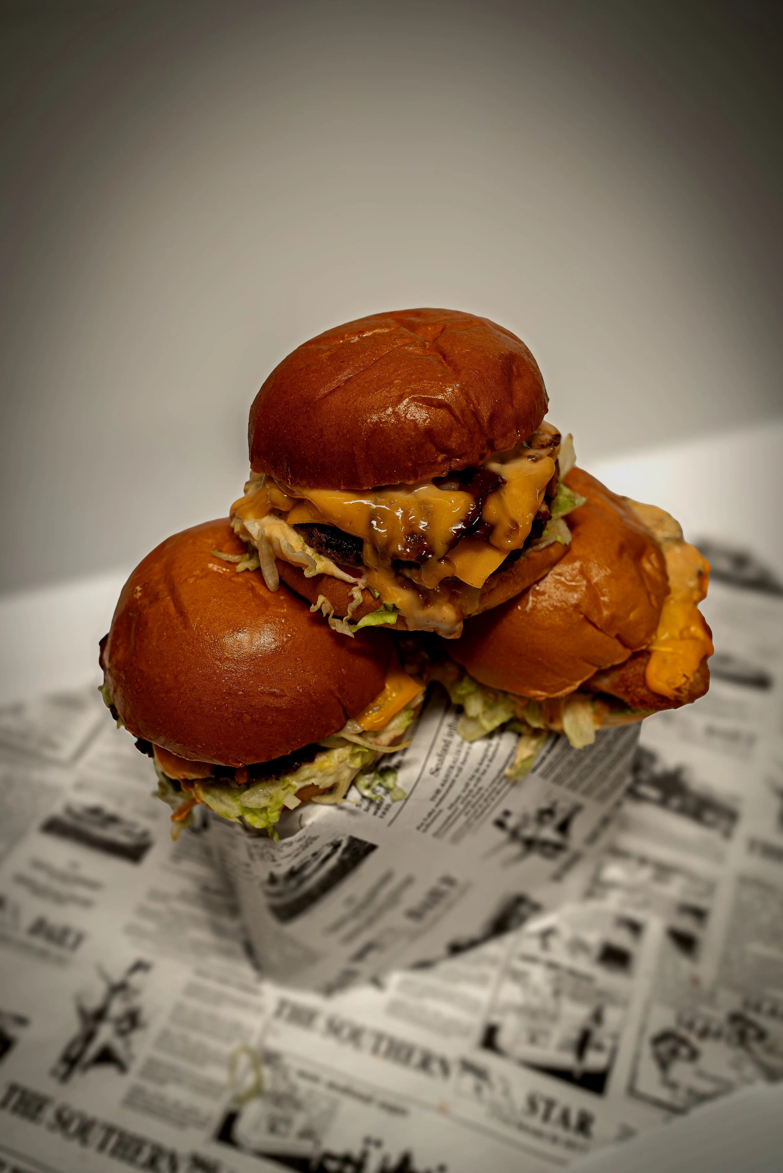 Three cheeseburgers stacked on top of each other with lettuce, shredded cheese, and sauces, placed on newspaper-lined surface.