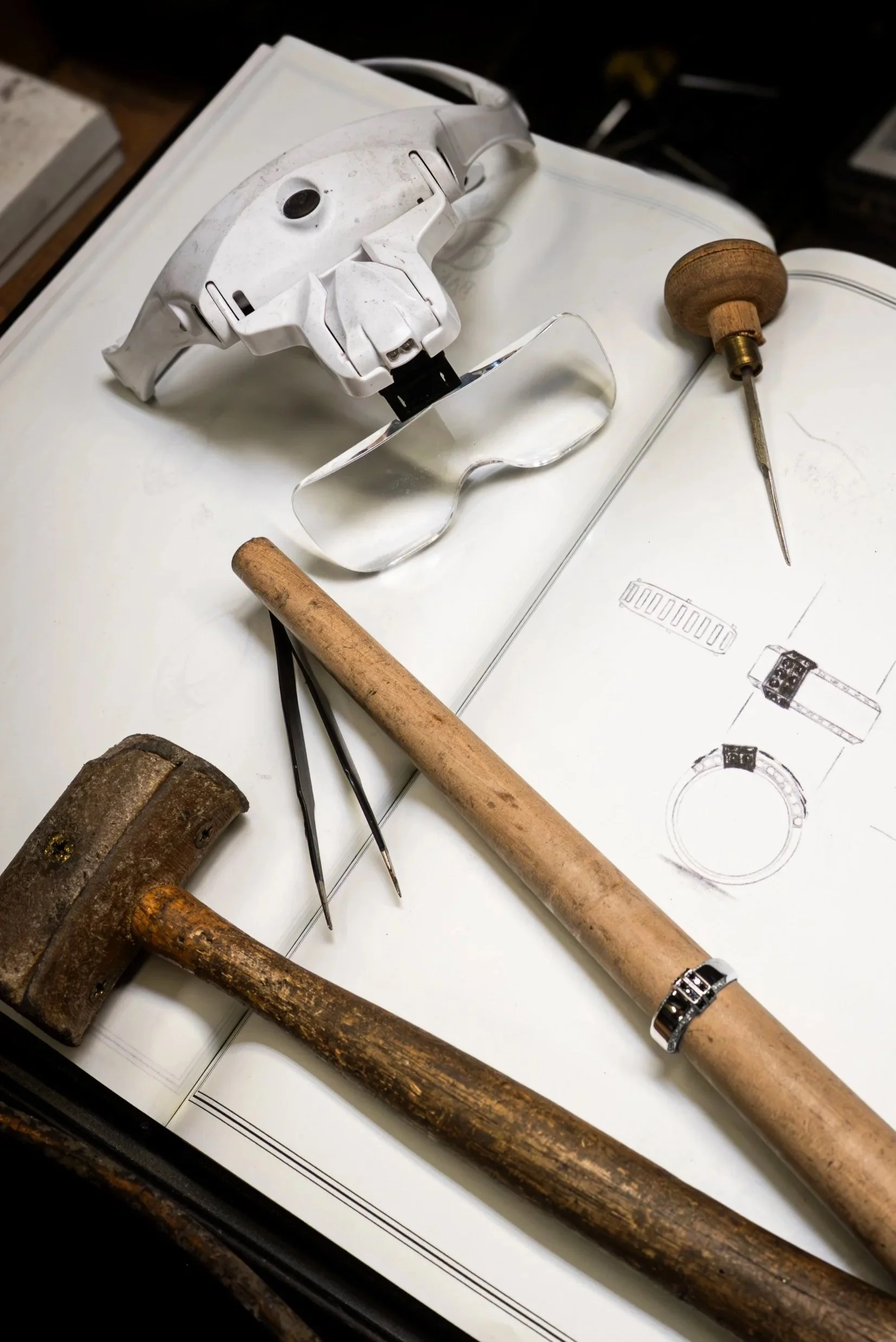 Tools including a hammer, screwdriver, punch, tweezers, protective glasses, and an accelerometer are placed on an open sheet with technical drawings of a watch.