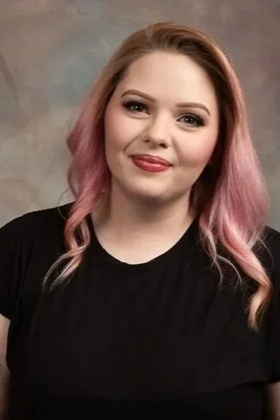 A woman with shoulder-length hair dyed pink and blonde, wearing a black shirt, smiling at the camera.