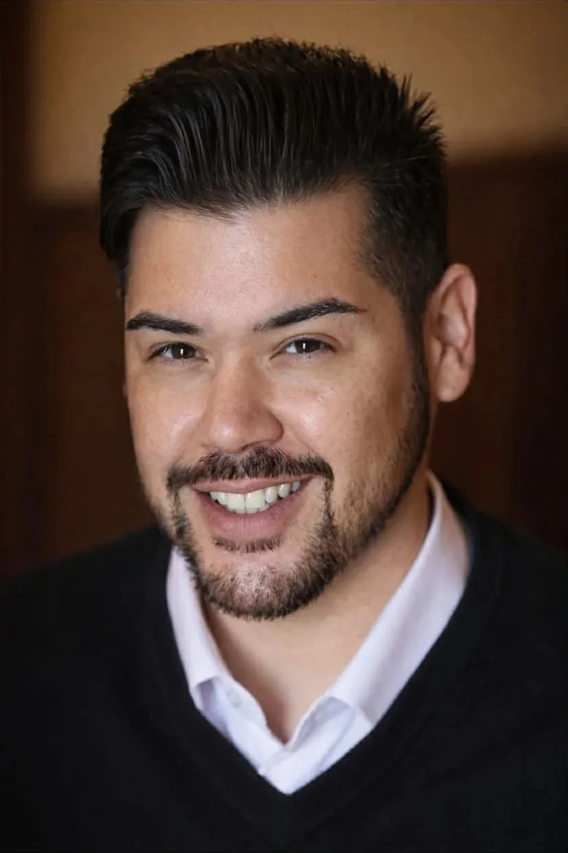 A man with dark hair, a beard, and a mustache, smiling, wearing a white shirt and black sweater, against a brown background.