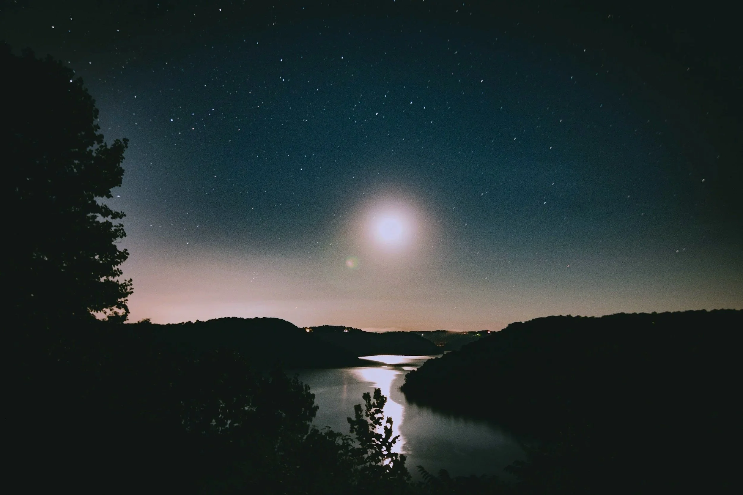 Night scene of a river flowing between hills, with a starry sky and a bright full moon overhead.