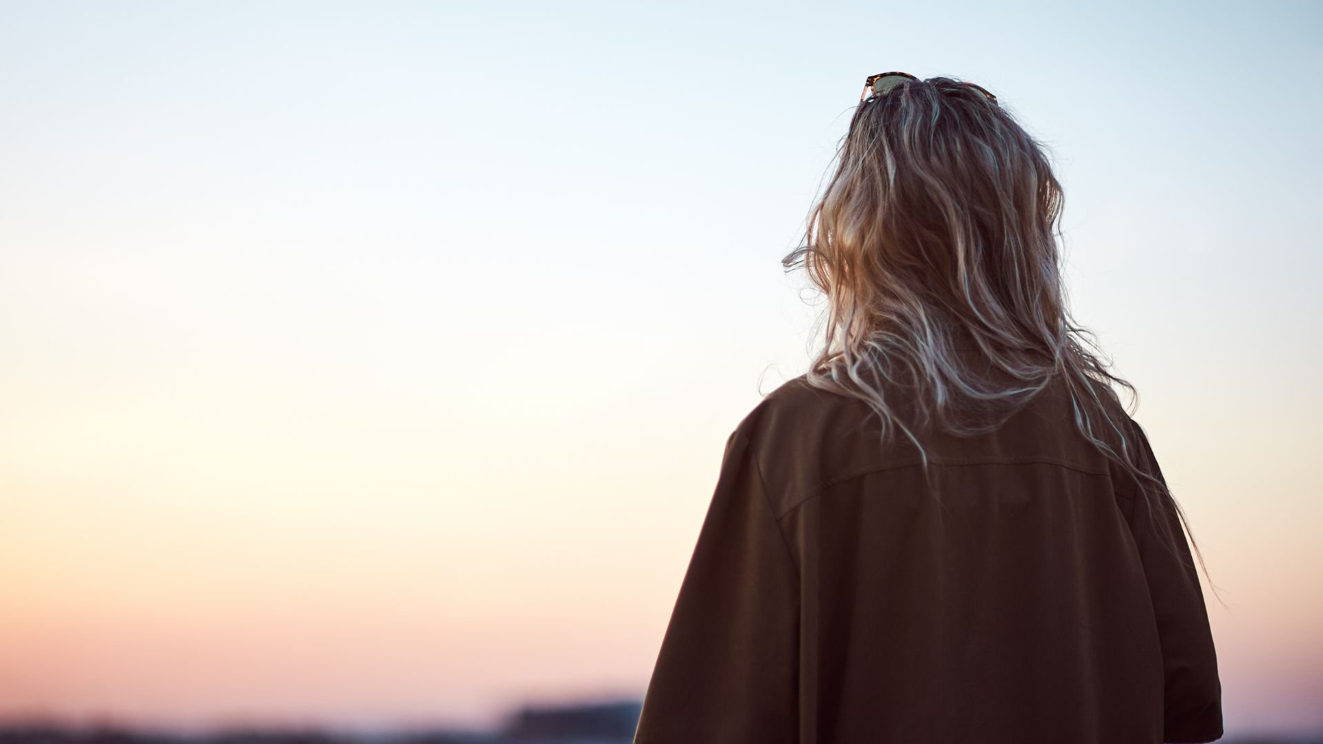 A woman with wavy hair and sunglasses resting on her head is seen from behind, looking at a sunset or sunrise sky.
