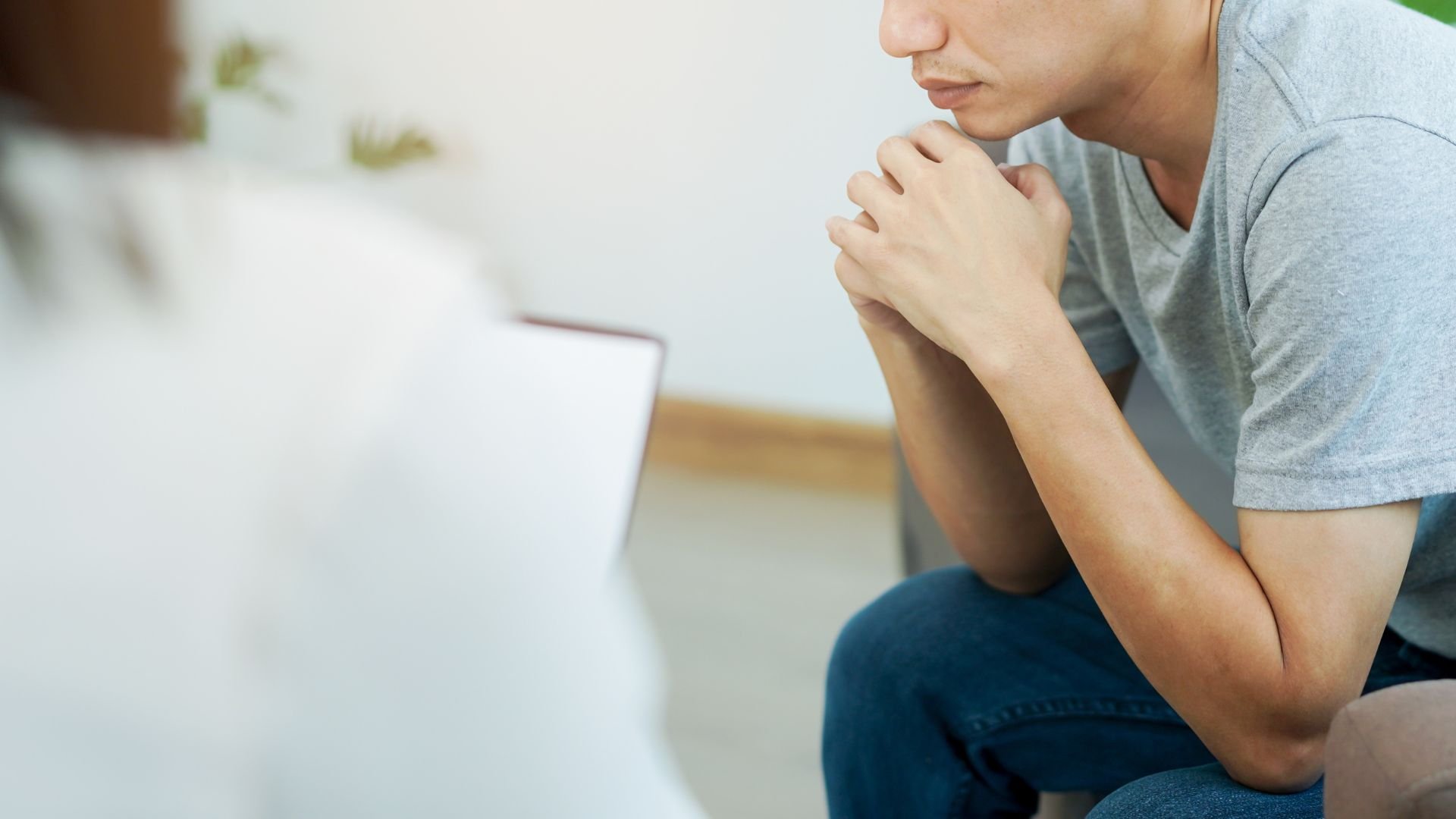 A man sitting indoors with his hands clasped near his chin, engaged in a serious conversation with another person whose shoulder is visible in the foreground.