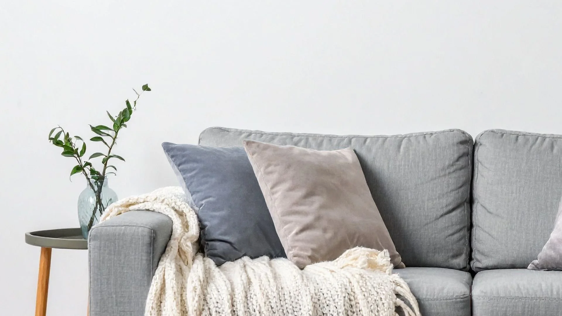 Living room scene with a light gray sofa adorned with three plush pillows in blue, beige, and light gray colors, a cozy cream-colored knitted throw blanket, and a small round side table with a wooden leg, holding a glass vase with green leafy branches, against a plain white wall.