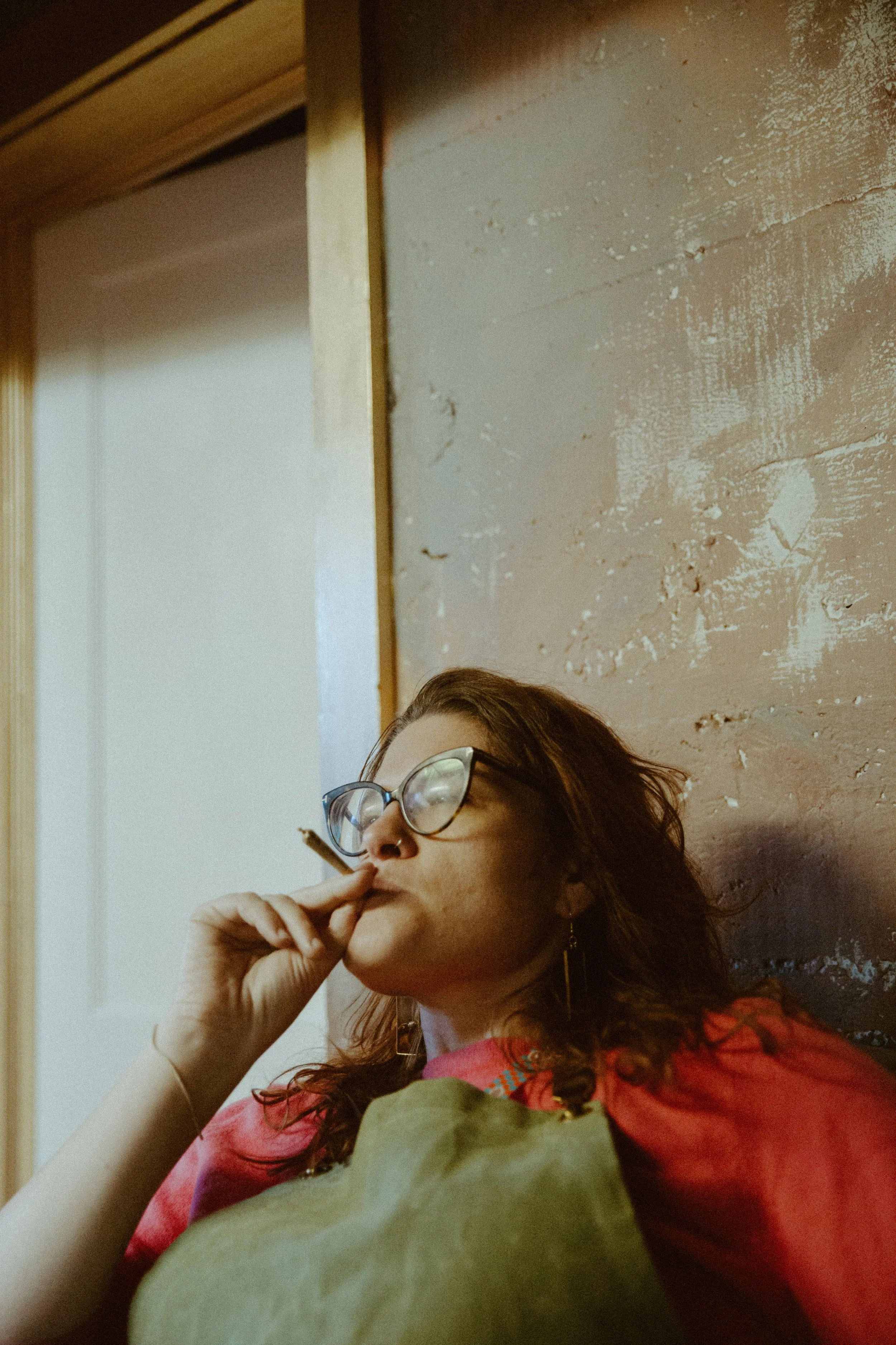 A woman with glasses and earrings, wearing a red and pink top, is sitting against a textured wall, smoking a cigarette.
