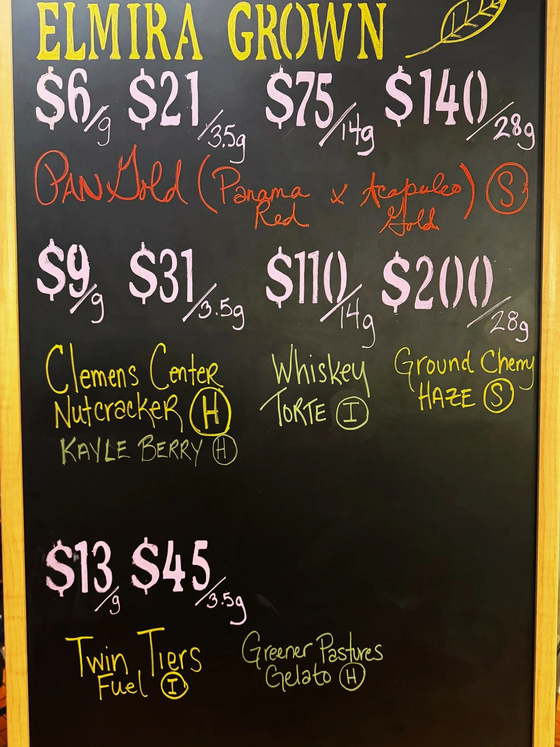 A blackboard menu displaying various cannabis products with prices, weights, and strains written in colorful chalk. The top section highlights Elmira Grown strains, including Band Old, with descriptions, and other strains like Clemens Center Nutcracker, Whiskey Torte, and Ground Chemy Haze at the bottom.