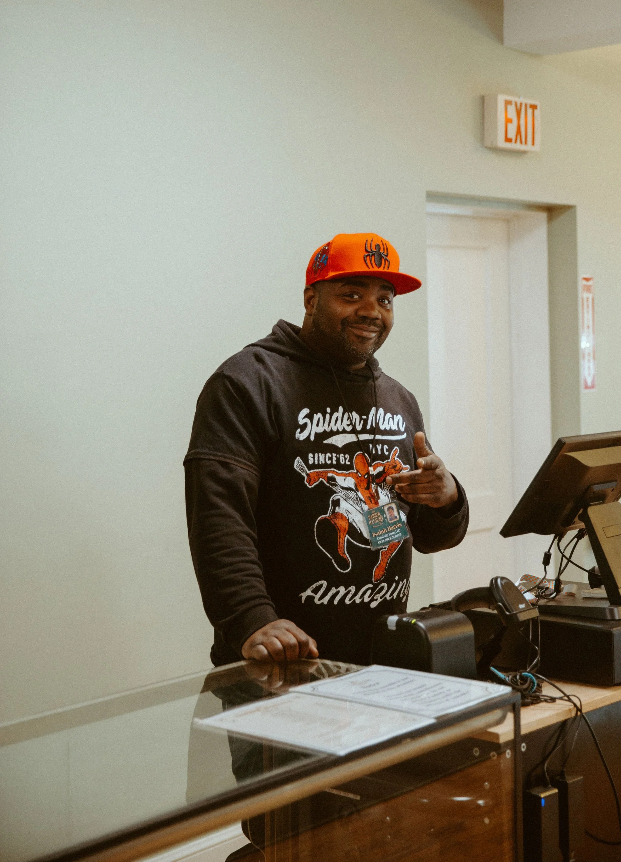 A man wearing a Spider-Man sweatshirt and a bright orange cap is posing at a reception desk. He is smiling and pointing at himself with his right hand. There is a computer and some papers on the desk, and an emergency exit sign is visible on the wall behind him.