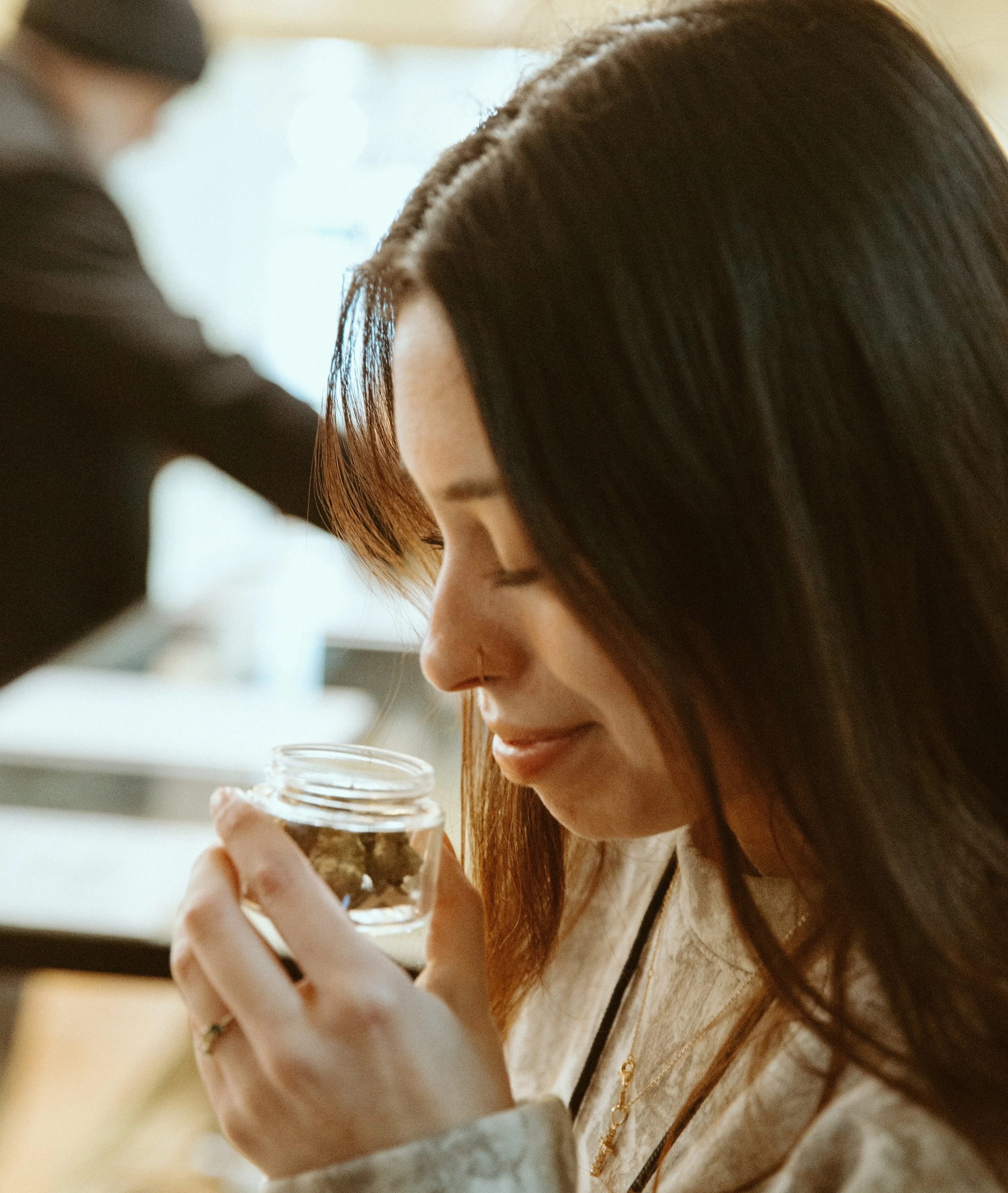 A woman with dark hair holding a small glass jar filled with cannabis buds, smiling with her eyes closed.