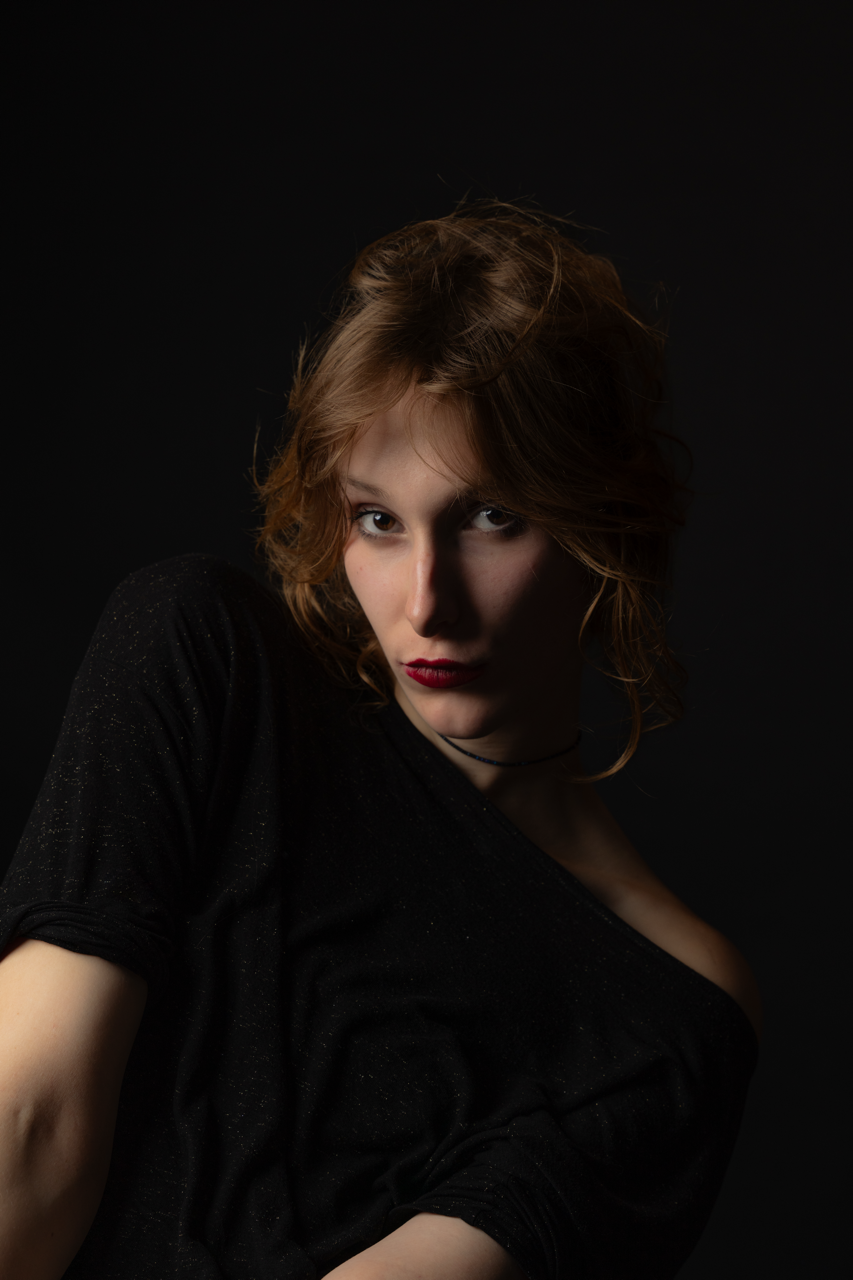 Portrait of a young woman with red hair wearing a black top against a dark background.