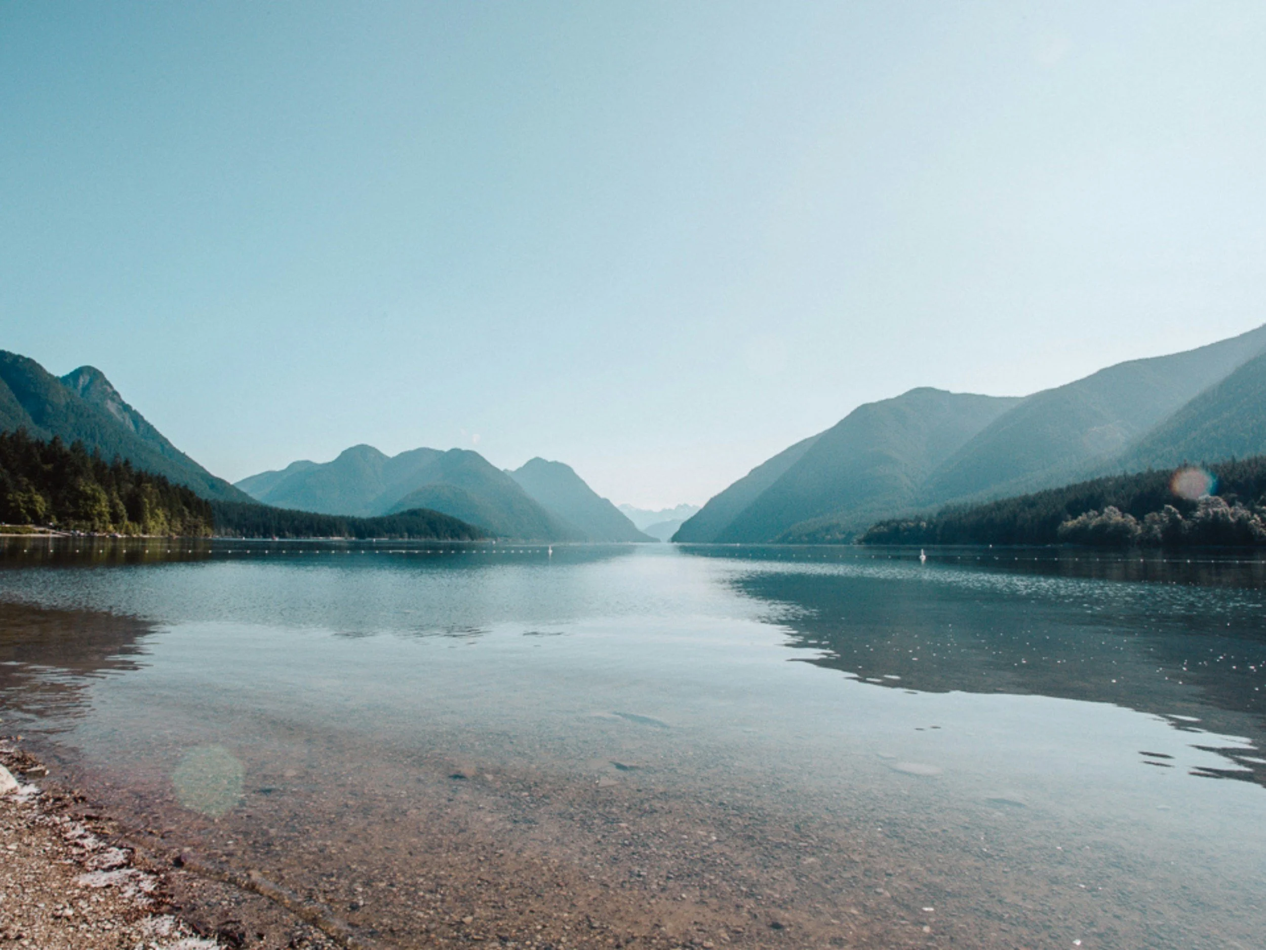 Calm lake surrounded by green mountains under clear sky, with a rocky shore in the foreground.