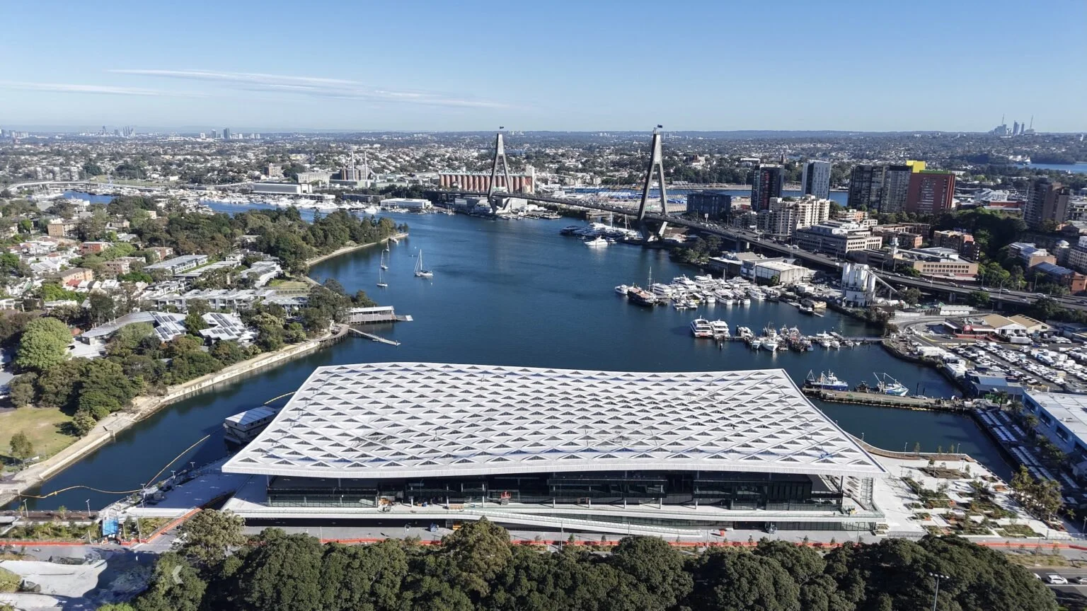 Aerial view of a city harbor with boats and sailboats, a bridge crossing the water, and a large modern building with a geometric patterned roof in the foreground.