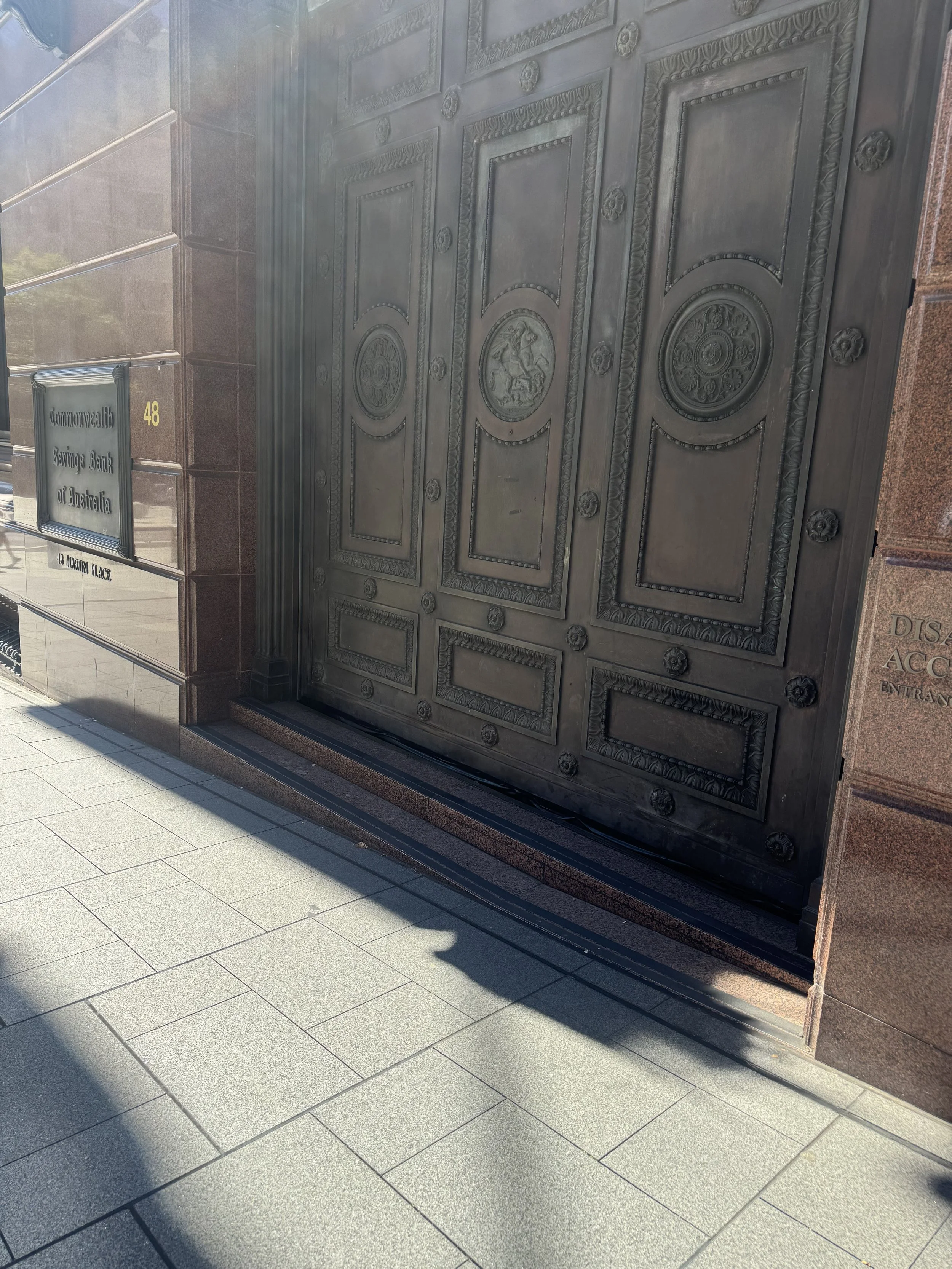 Large ornate metal door with intricate designs and carvings, part of a brick building, with a sidewalk and shadows in the foreground.