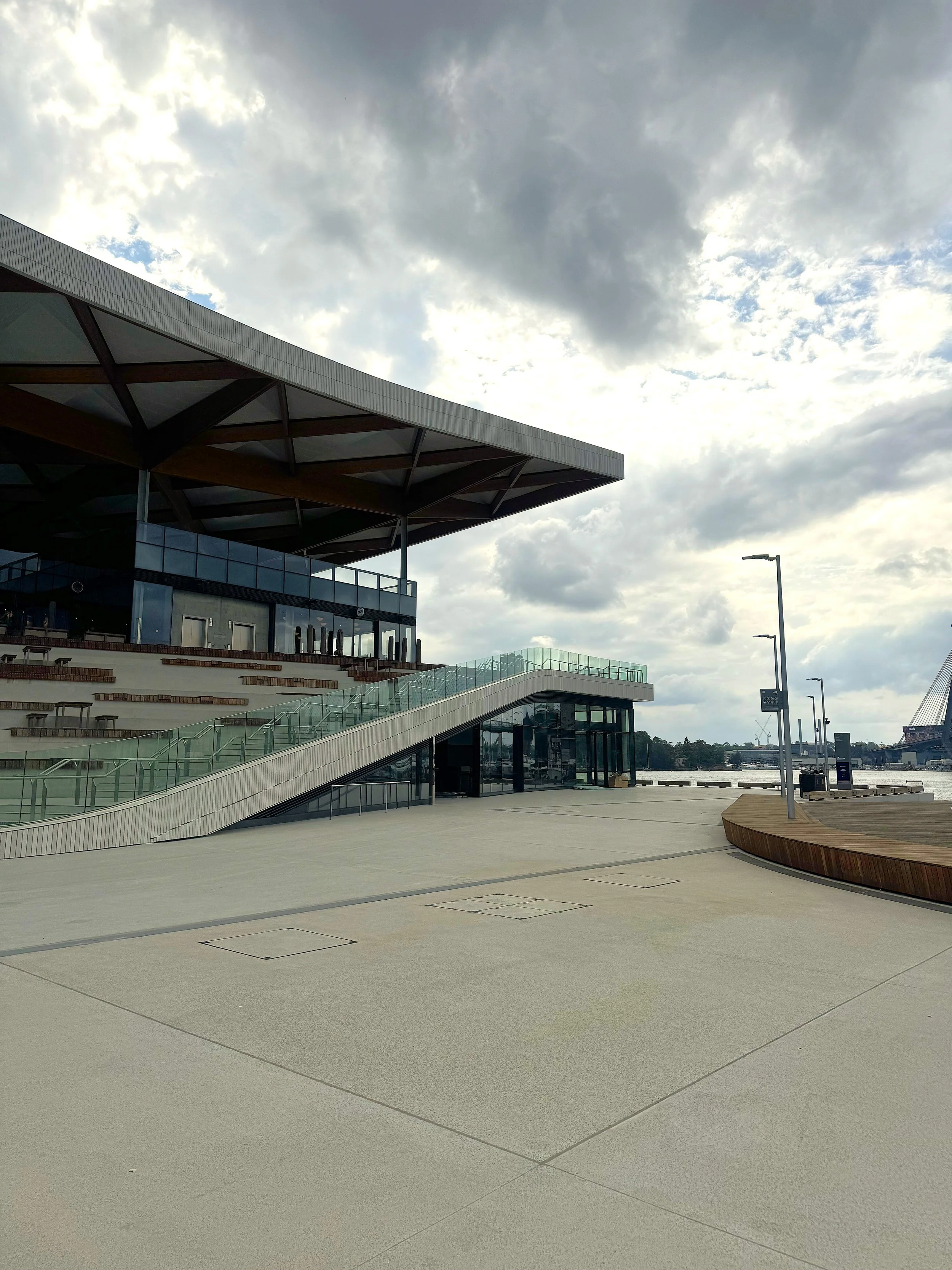 Modern waterfront building with glass walls and outdoor seating area under a cloudy sky.