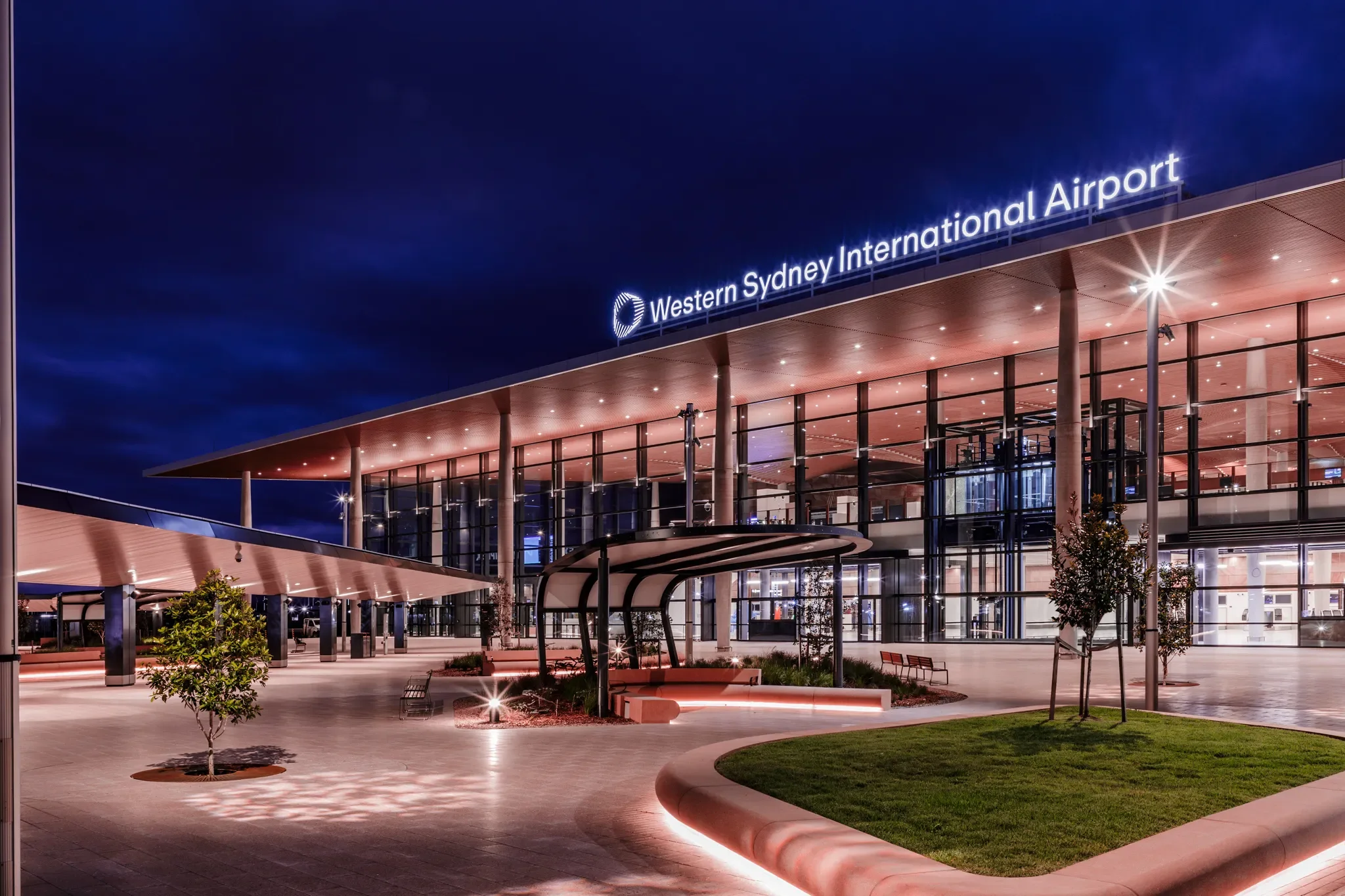 Exterior of Western Sydney International Airport at night with illuminated signage, glass facade, landscaped planters, and seating area