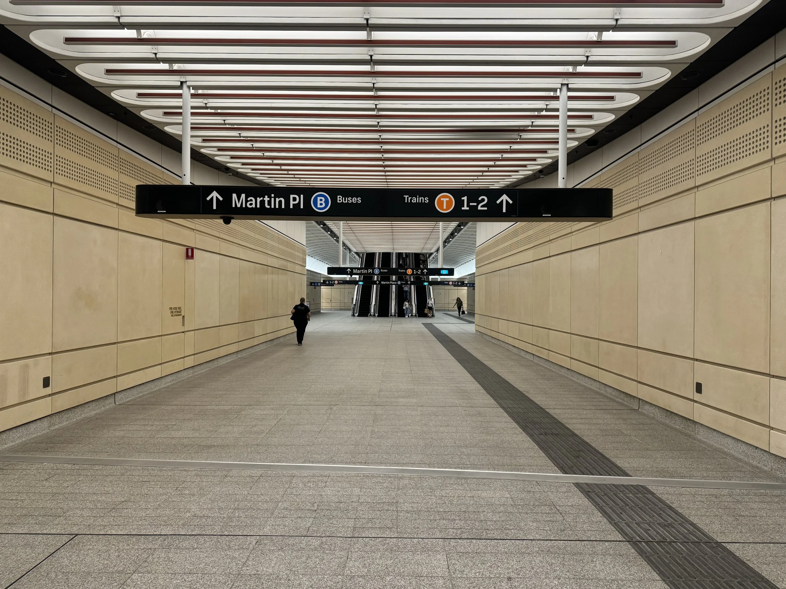 Interior of a modern train station with beige walls, a high ceiling with white and red accents, and a black overhead sign indicating directions to Martin Plaza, buses, and trains. There are escalators in the background with a few people walking.