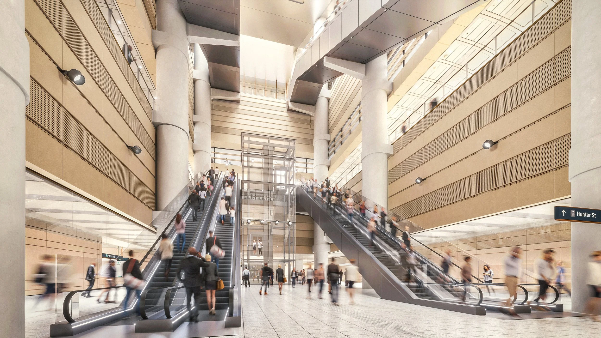 Interior of a modern train station with multiple escalators and people walking, some ascending and descending the escalators, in a spacious area with high ceilings and beige and gray walls.