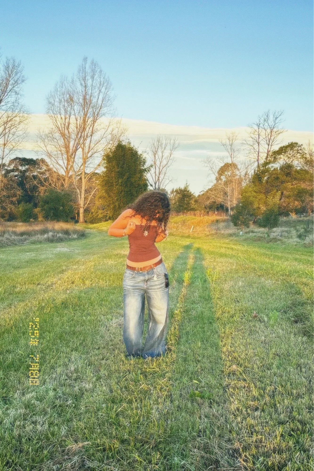 A woman with curly hair looking down, standing on a grassy field during sunset with trees in the background.