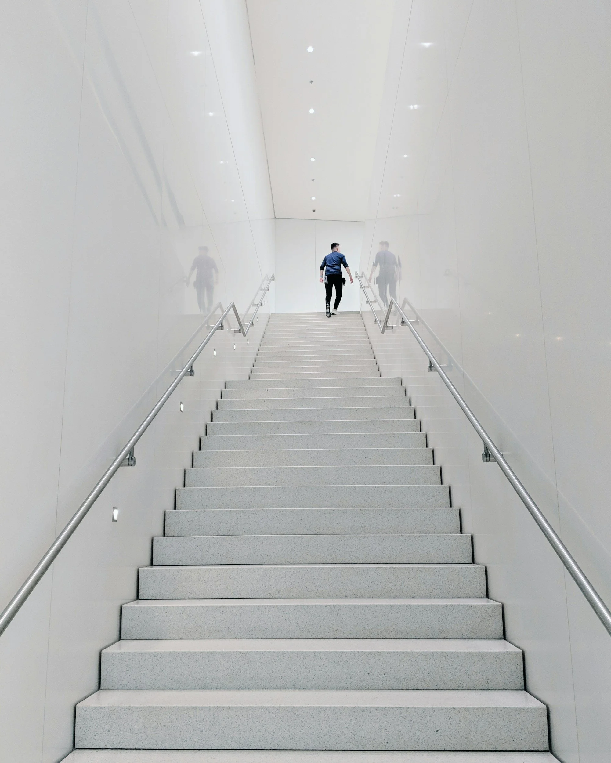 A man in a blue shirt and dark pants walking up a staircase in a modern, minimalistic building with white walls and stainless steel railings.
