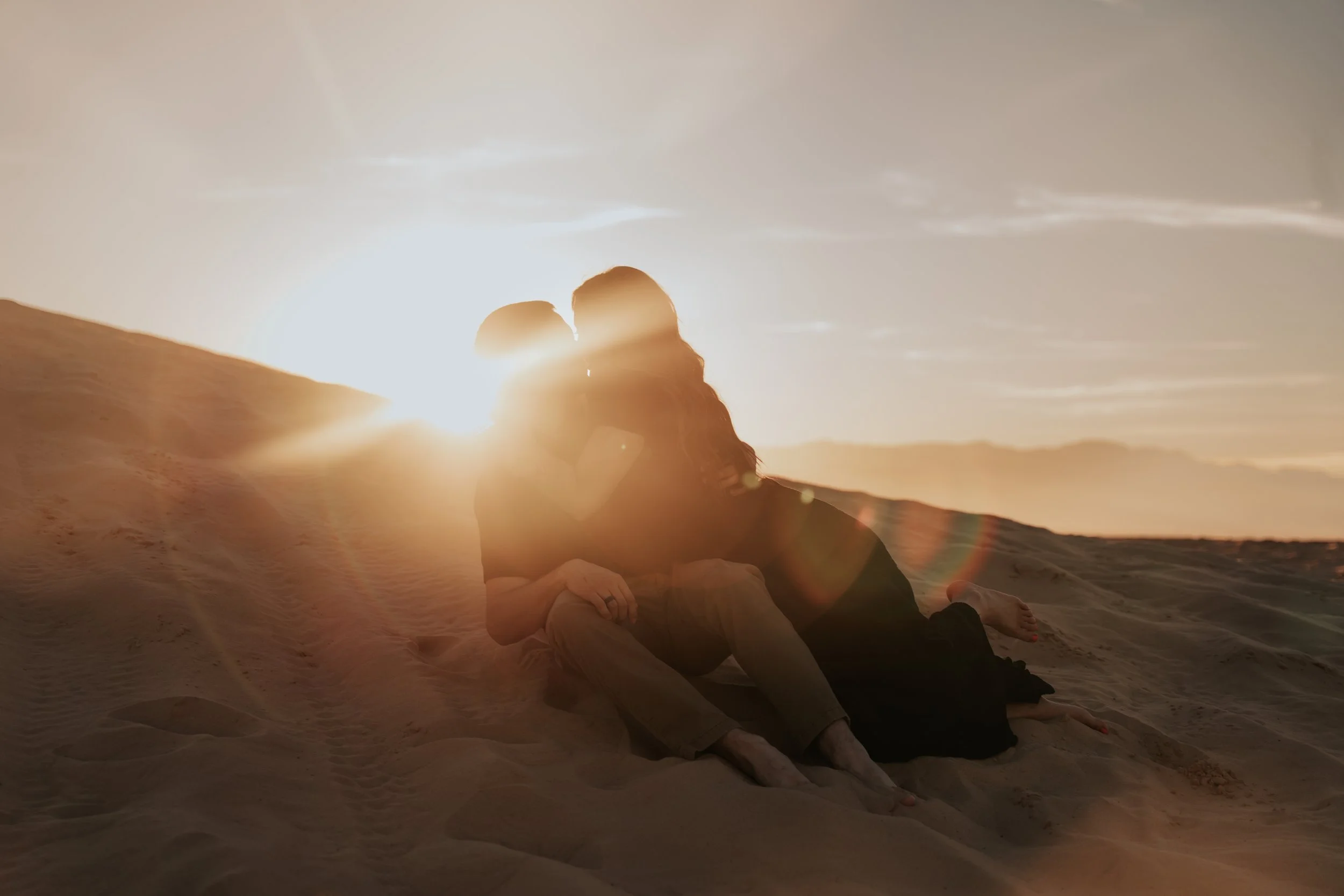 A couple sitting on sand dunes during sunset, embracing and kissing, with sunlight creating a warm glow and lens flare.