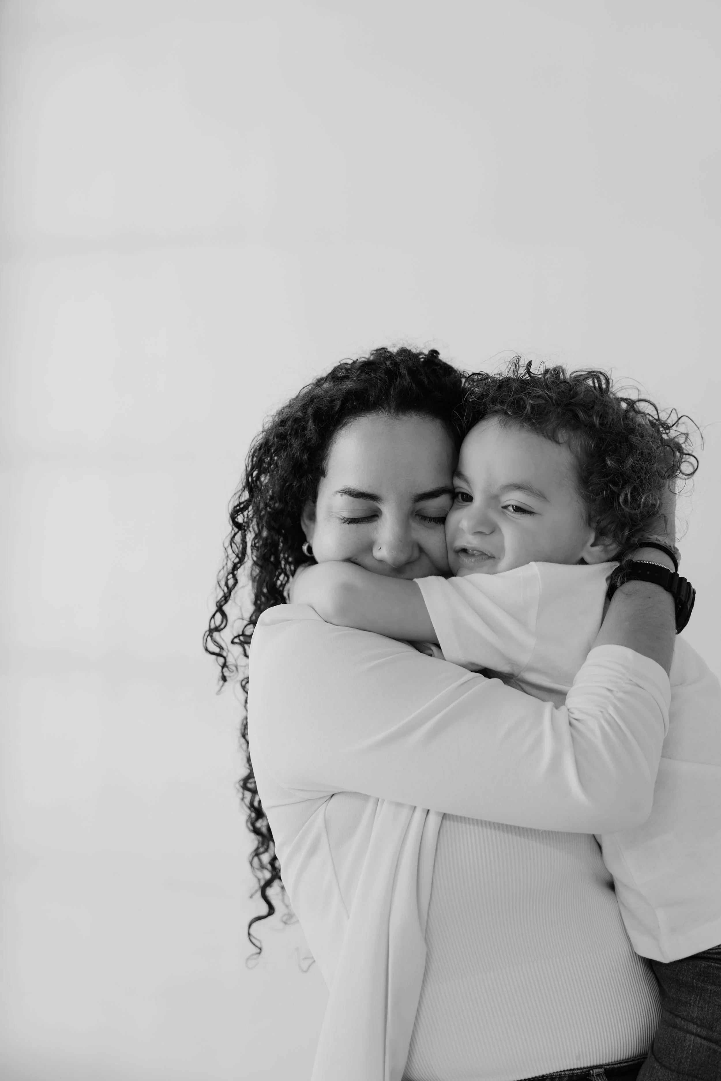 A woman with curly hair hugging a young boy, both with eyes closed, in a heartfelt embrace.