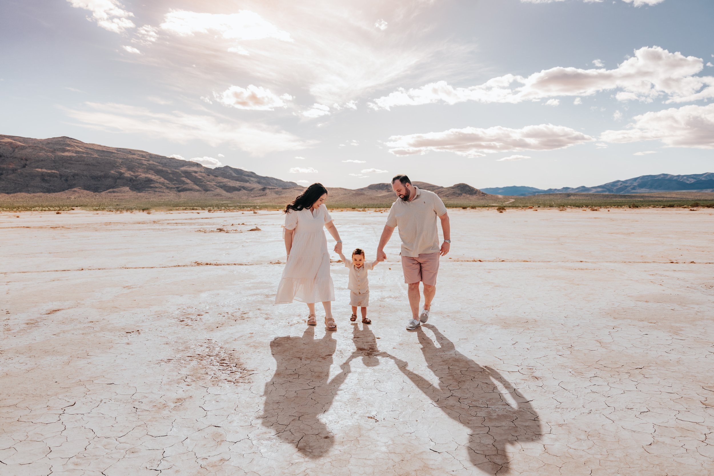 A family of three, a woman, a man, and a small child, holding hands and walking on a dry, cracked salt flat with mountains and a partly cloudy sky in the background.