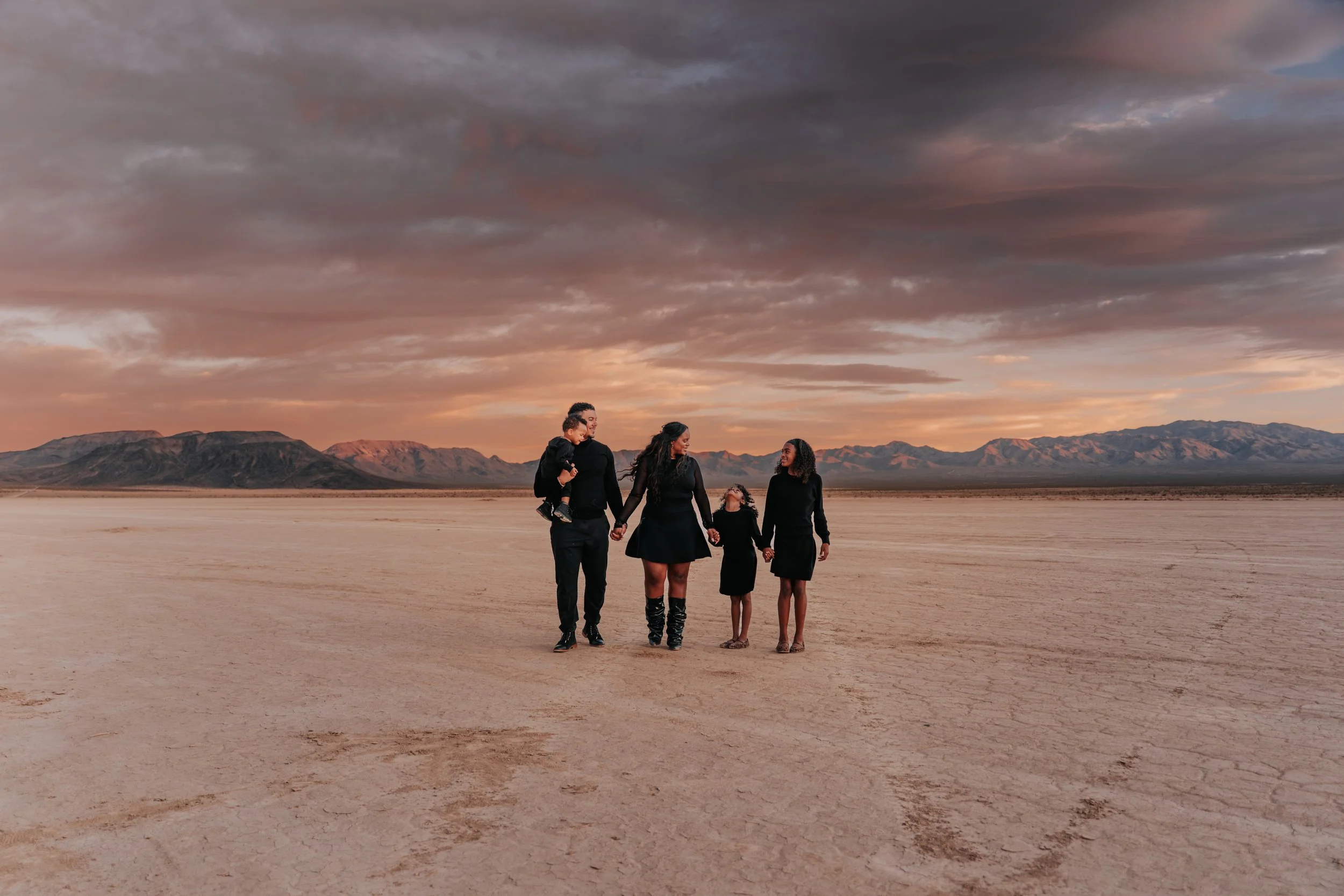 A family of five dressed in black walking in a dry, desert landscape with mountains in the background during sunset.