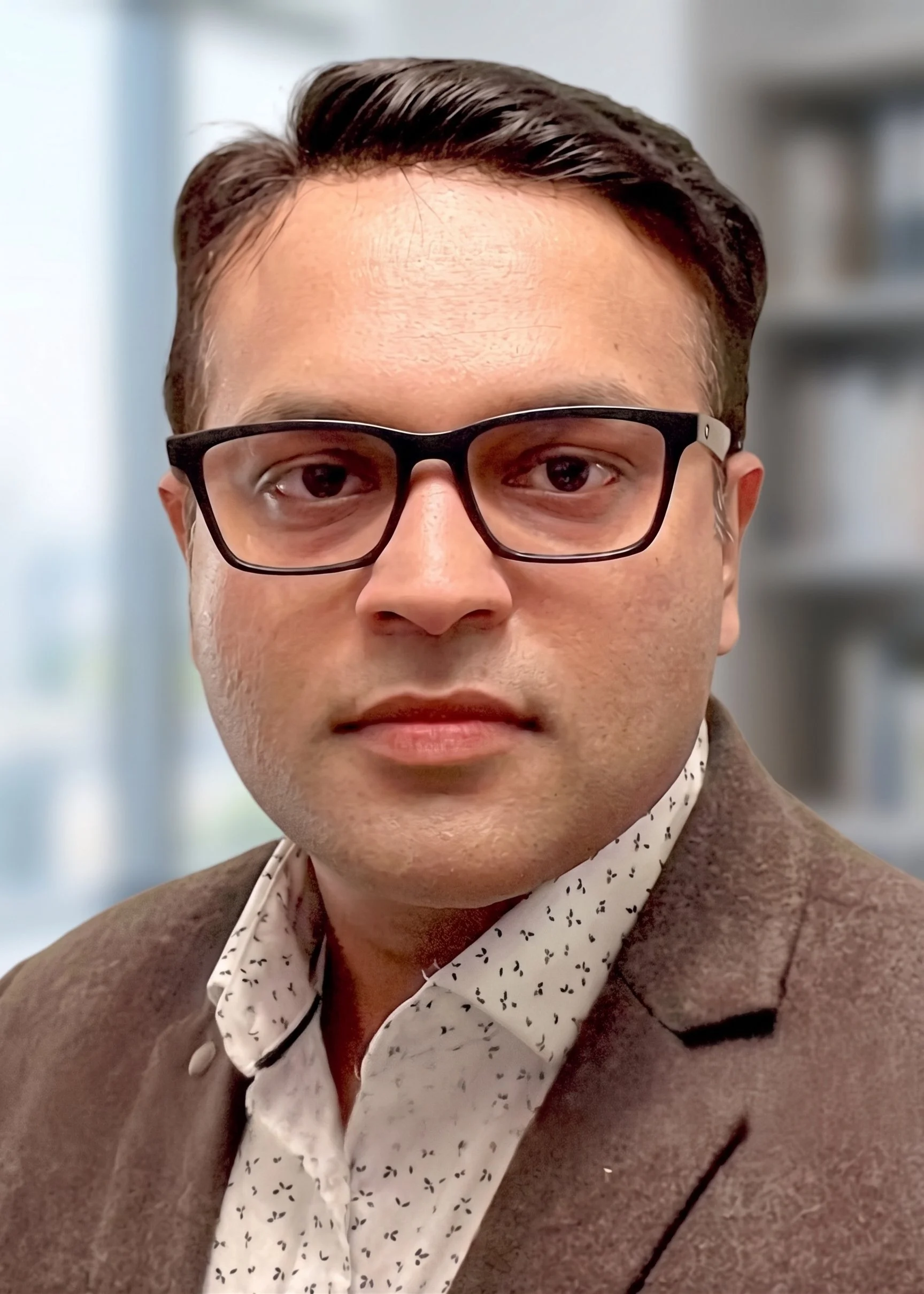 Close-up portrait of a man wearing black glasses, a brown blazer, and a white shirt with black pattern, in an indoor setting.