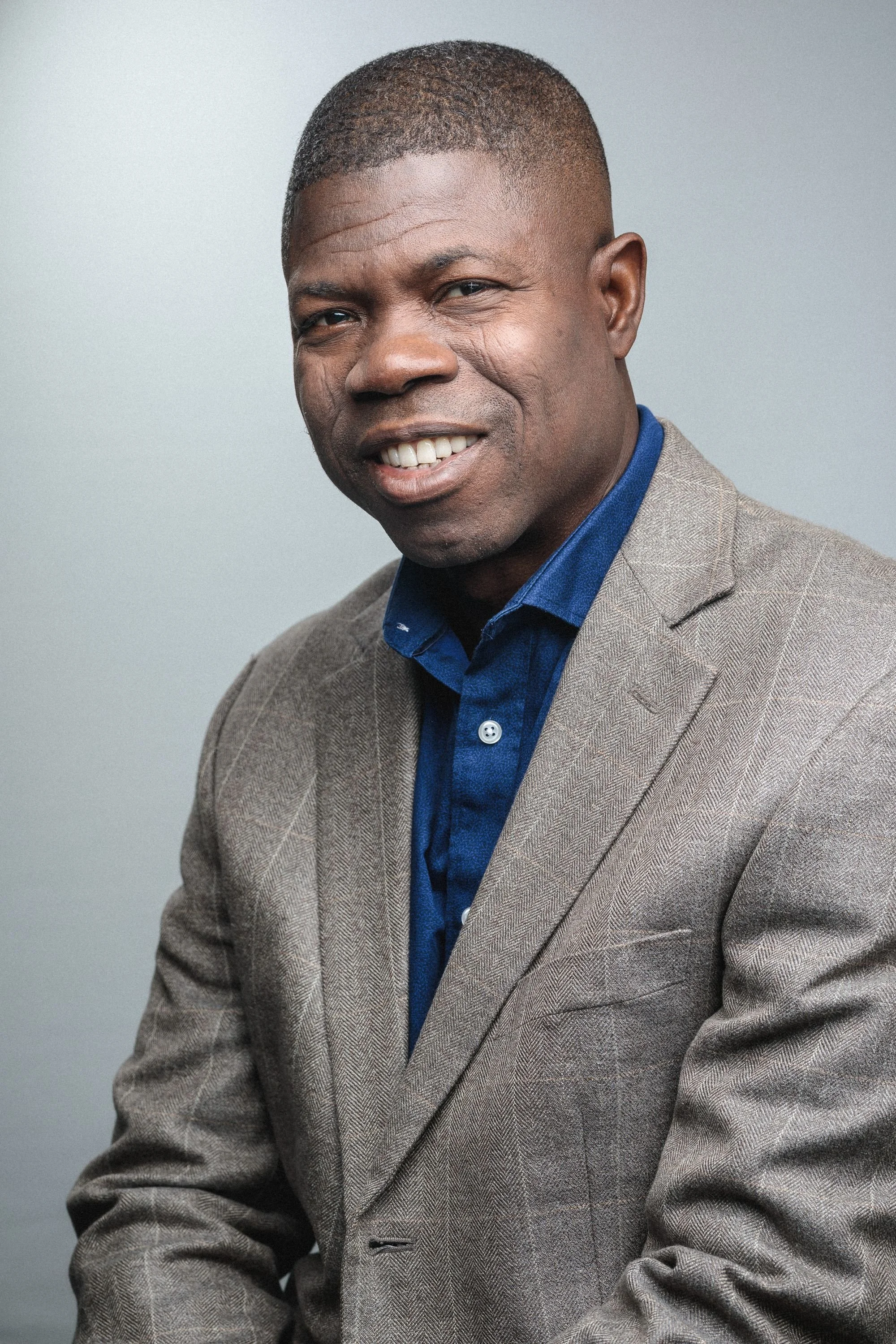A portrait of a smiling African-American man with short hair, wearing a beige plaid blazer and a blue collared shirt, posing against a neutral gray background.