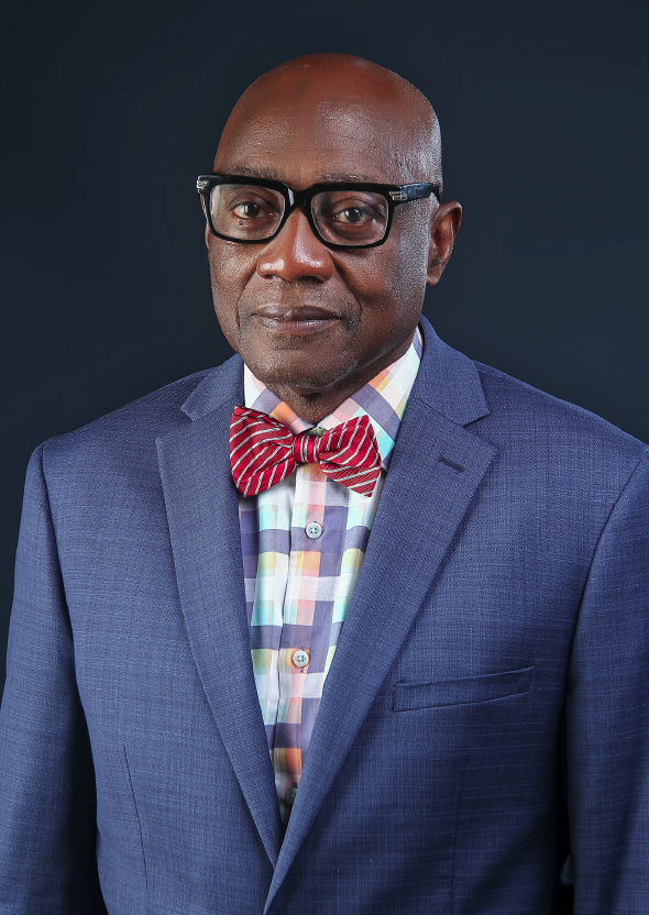 A man wearing black glasses, a blue suit, a colorful checkered shirt, and a red striped bow tie against a dark background.
