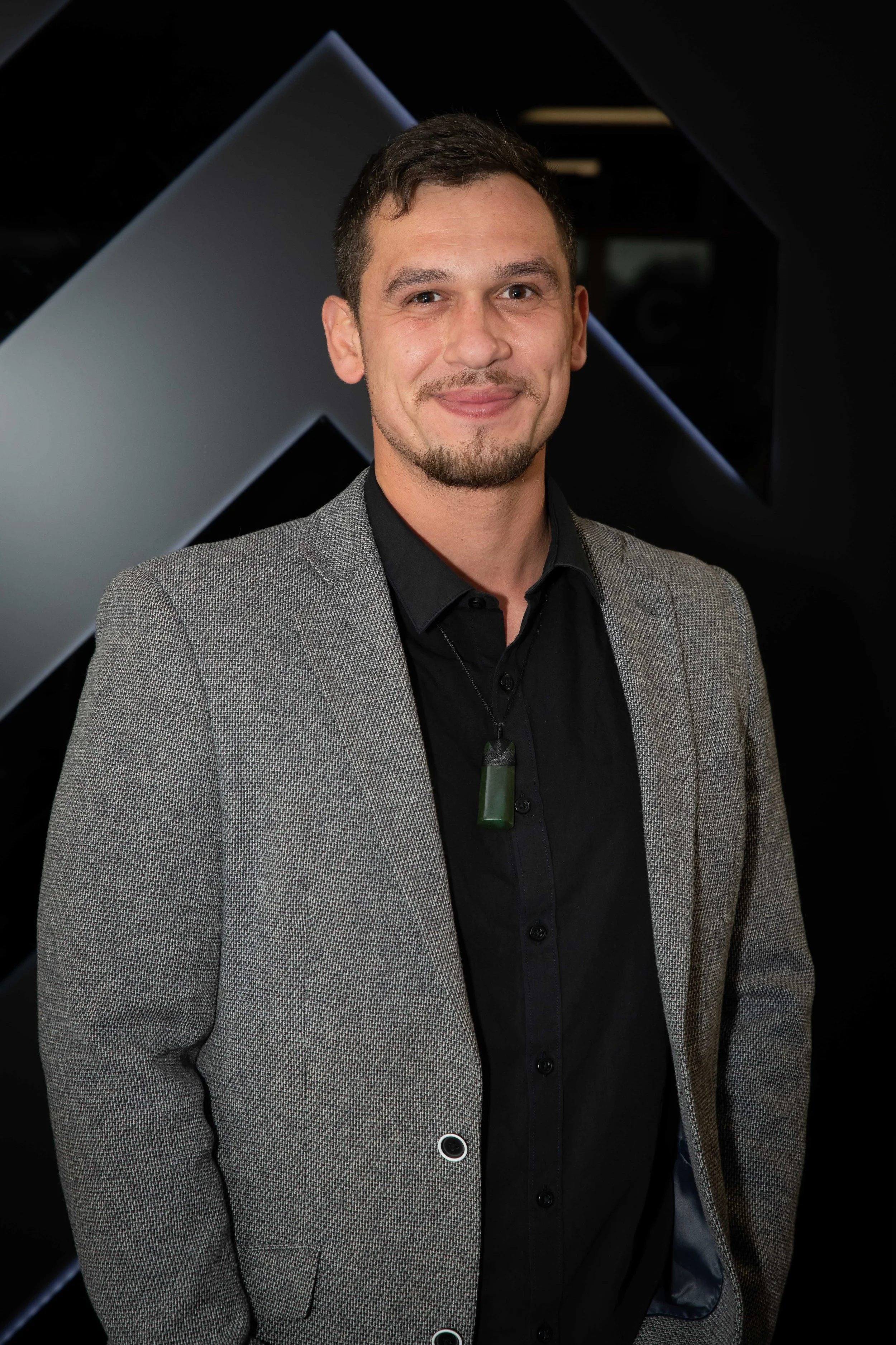 A man with short dark hair, a goatee, and a mustache posing for a photo. He is wearing a gray blazer over a black shirt and a pendant necklace with a green rectangular stone. The background features dark, geometric shapes.
