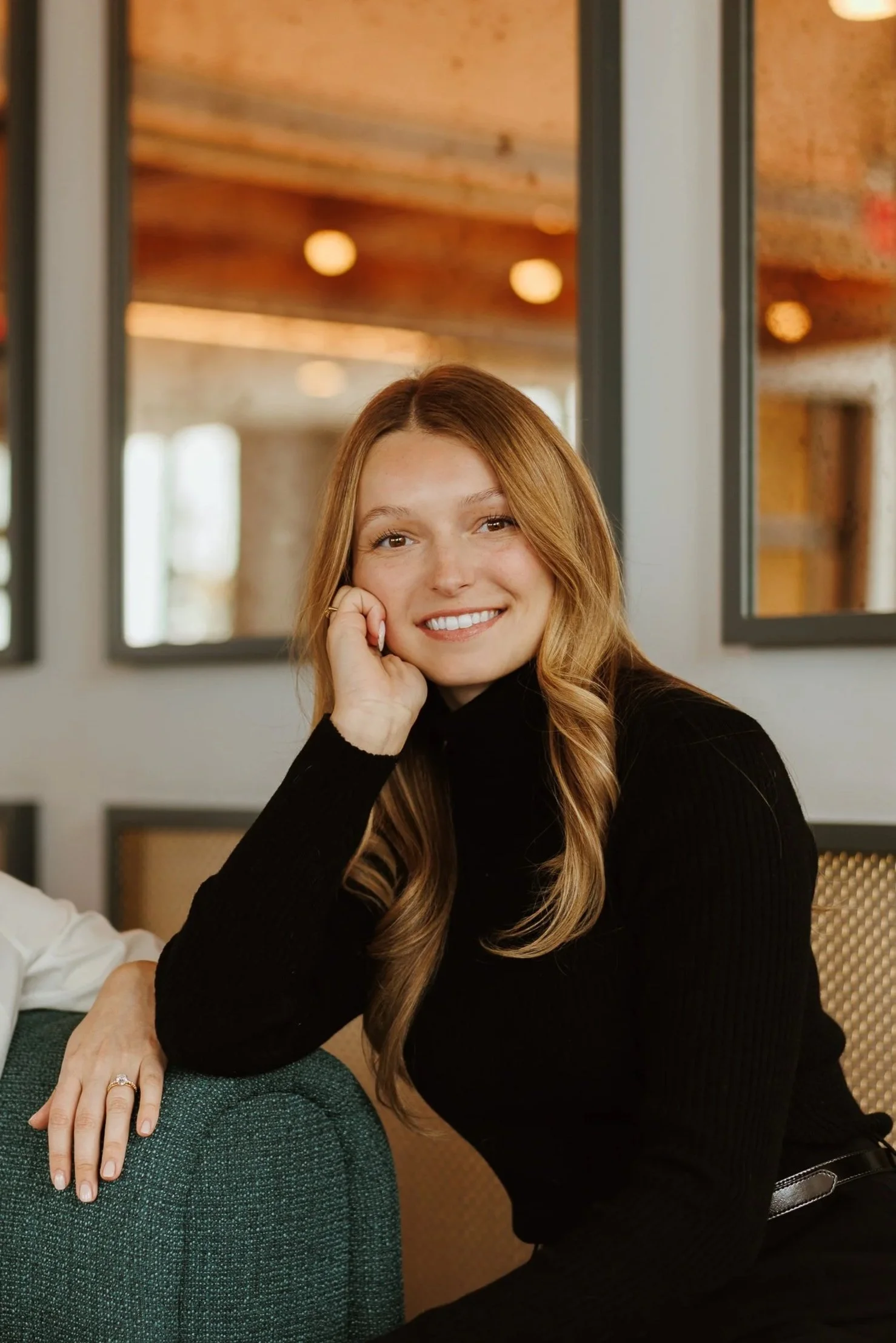 A woman with long blonde hair, smiling, resting her chin on her hand, sitting indoors with a woman beside her, wearing a black top, and a room with large windows and warm lighting in the background.