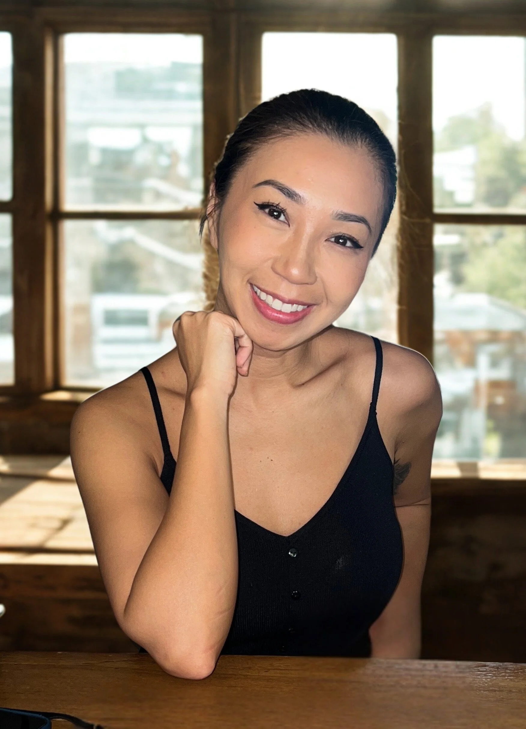 A smiling woman with dark hair pulled back, wearing a black spaghetti strap top, sitting at a wooden table in front of a window with a blurred outdoor scene.