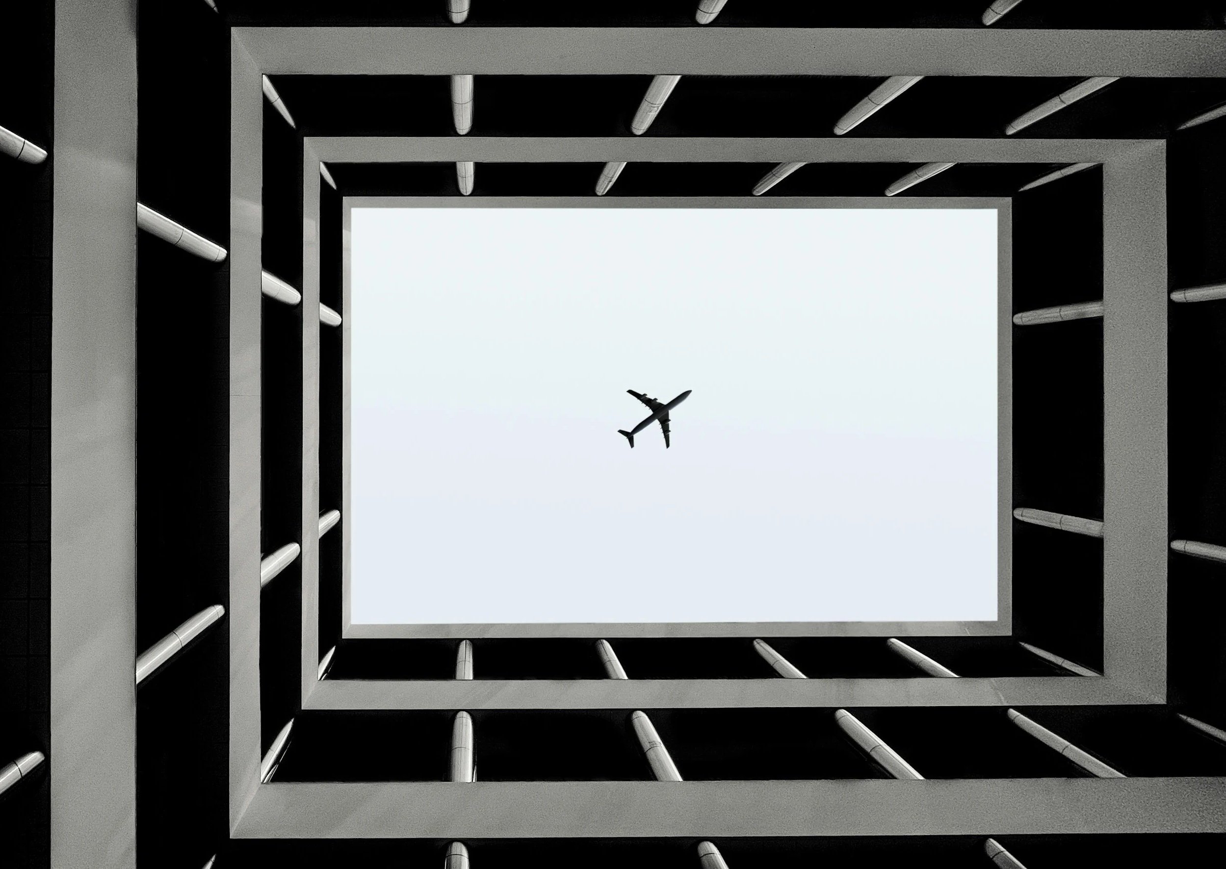 Looking up through the open courtyard of a building with layered square frames and bars, with a plane flying in the sky above.