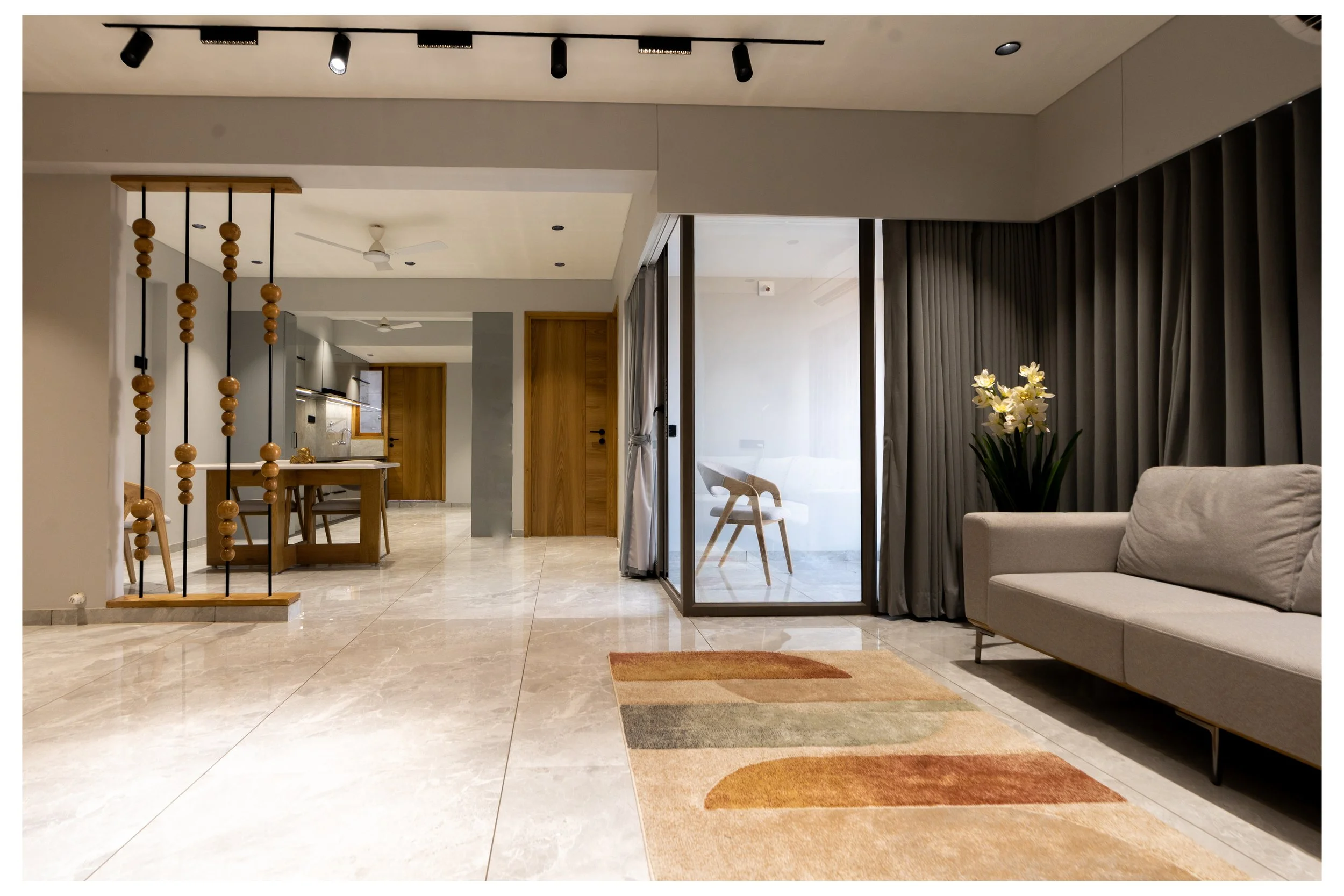 Modern living room with beige marble flooring, a colorful area rug, a light gray sofa, a vase with white flowers, and a glass-enclosed balcony with a wooden chair. The space includes a dining area in the background, a wooden door, and a partition wit