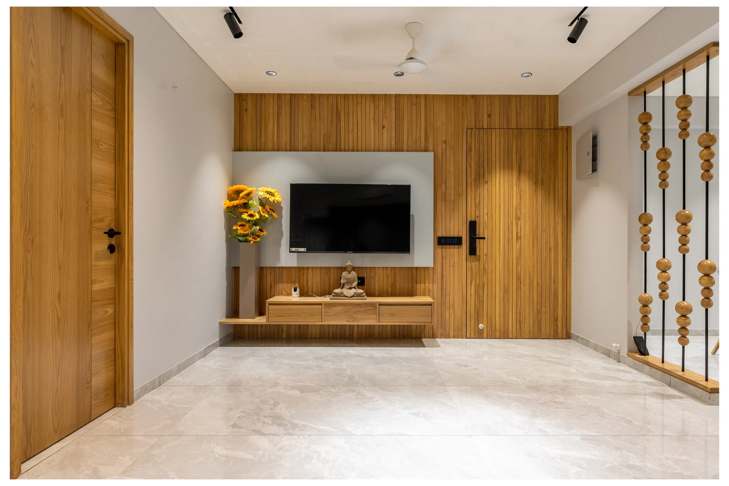 Modern interior living room with a flat-screen TV mounted on a wood-paneled accent wall, a floating wooden shelf with a Buddha statue, a vase with sunflowers, and a small decorative object, beige marble flooring, and a wooden door on the left. Partia