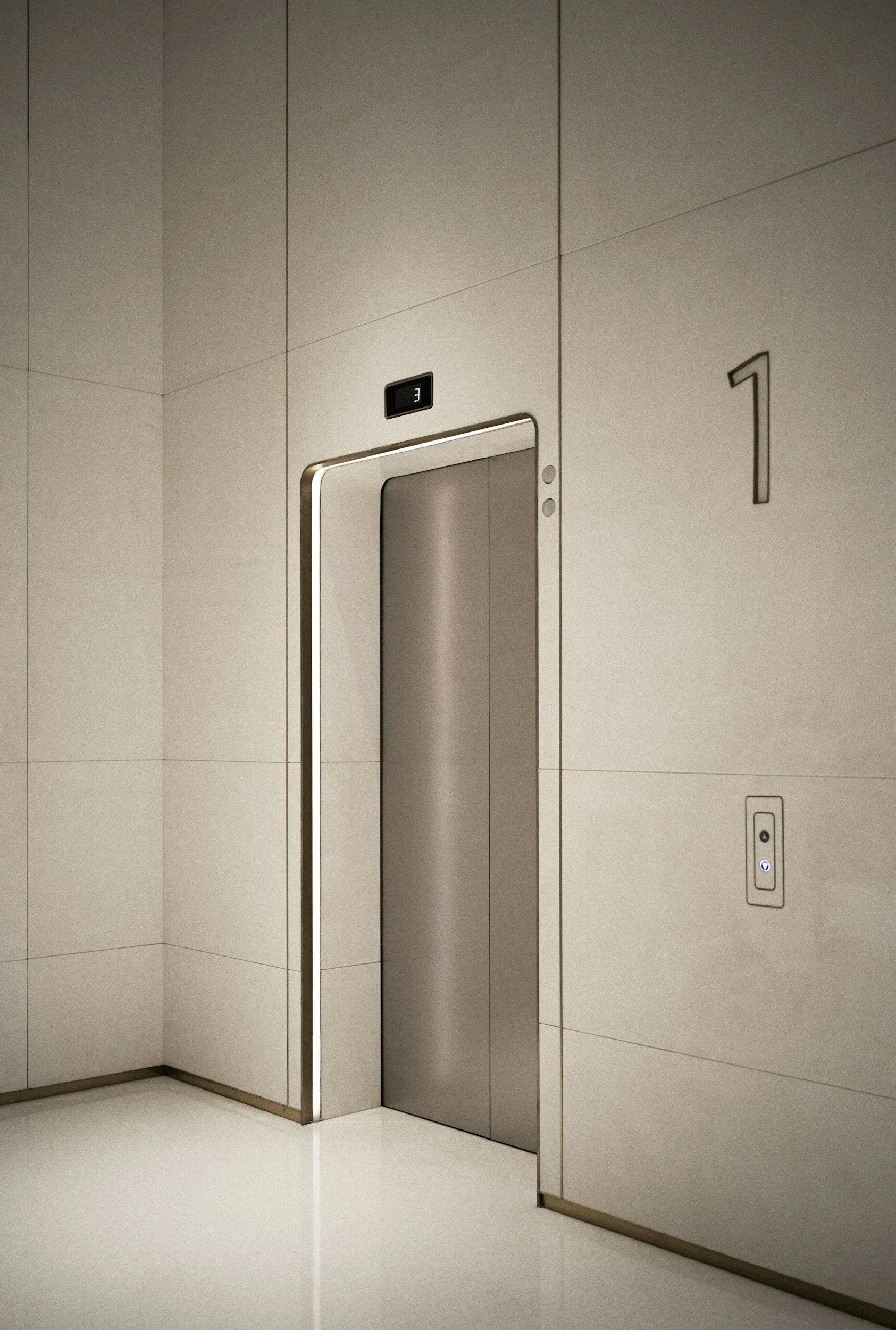 Modern elevator with closed doors in a minimalist building with a digital floor display showing the number 3, adjacent to a large number 1 on the wall.