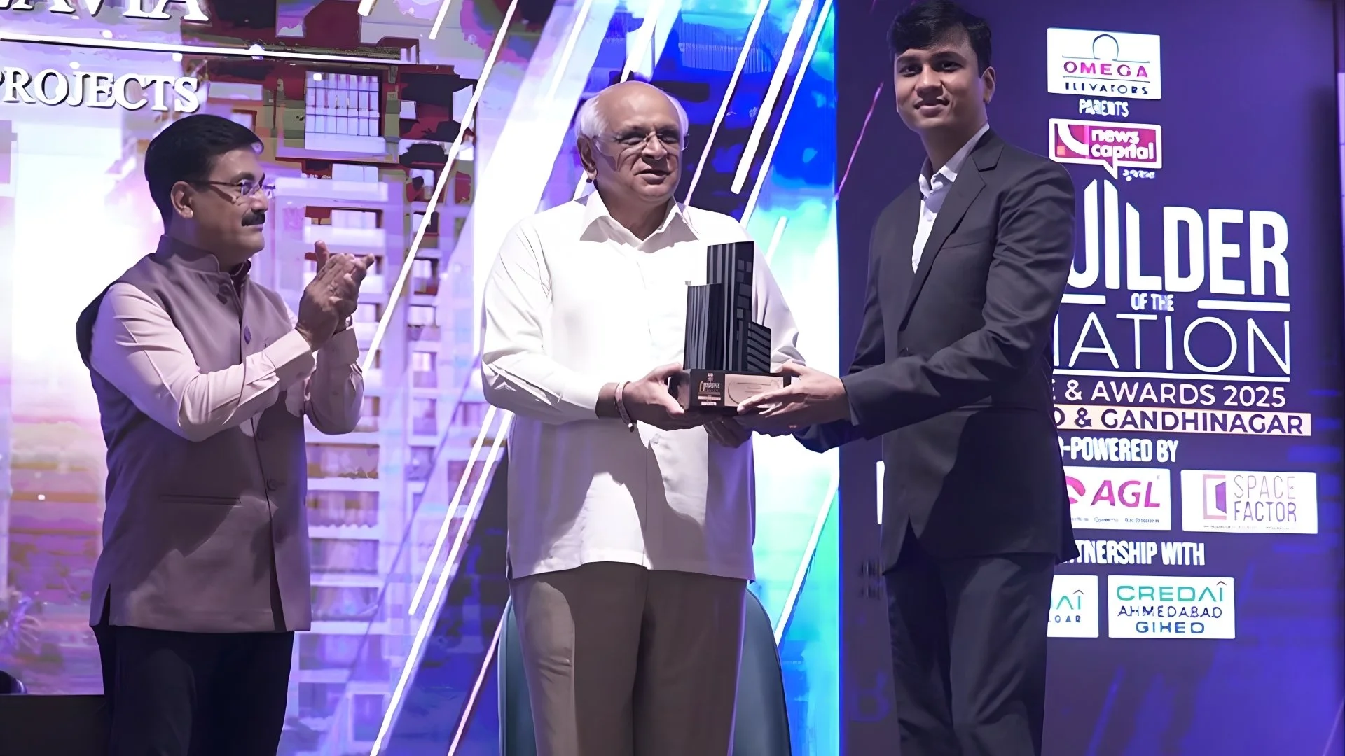 An award ceremony where a young man in a black suit is receiving a trophy from an older man in a white shirt. A man with glasses and a traditional outfit is clapping in the background. The backdrop shows logos and text about the event, including 'Builder of the Nation 2025' and city names.
