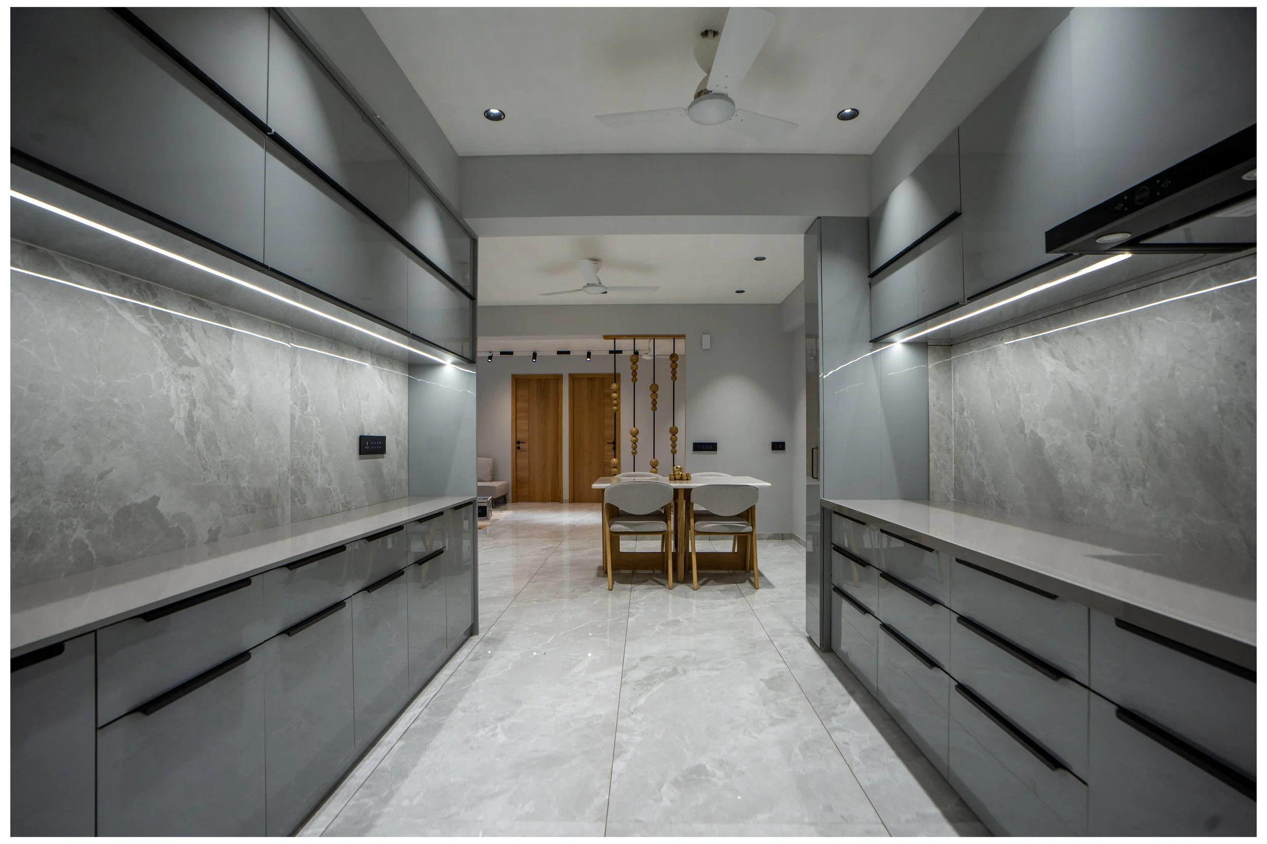 Modern kitchen with gray cabinets and marble countertops, view from the entrance looking toward a dining area with a wooden table and white chairs, ceiling fans, and wooden doors in the background.