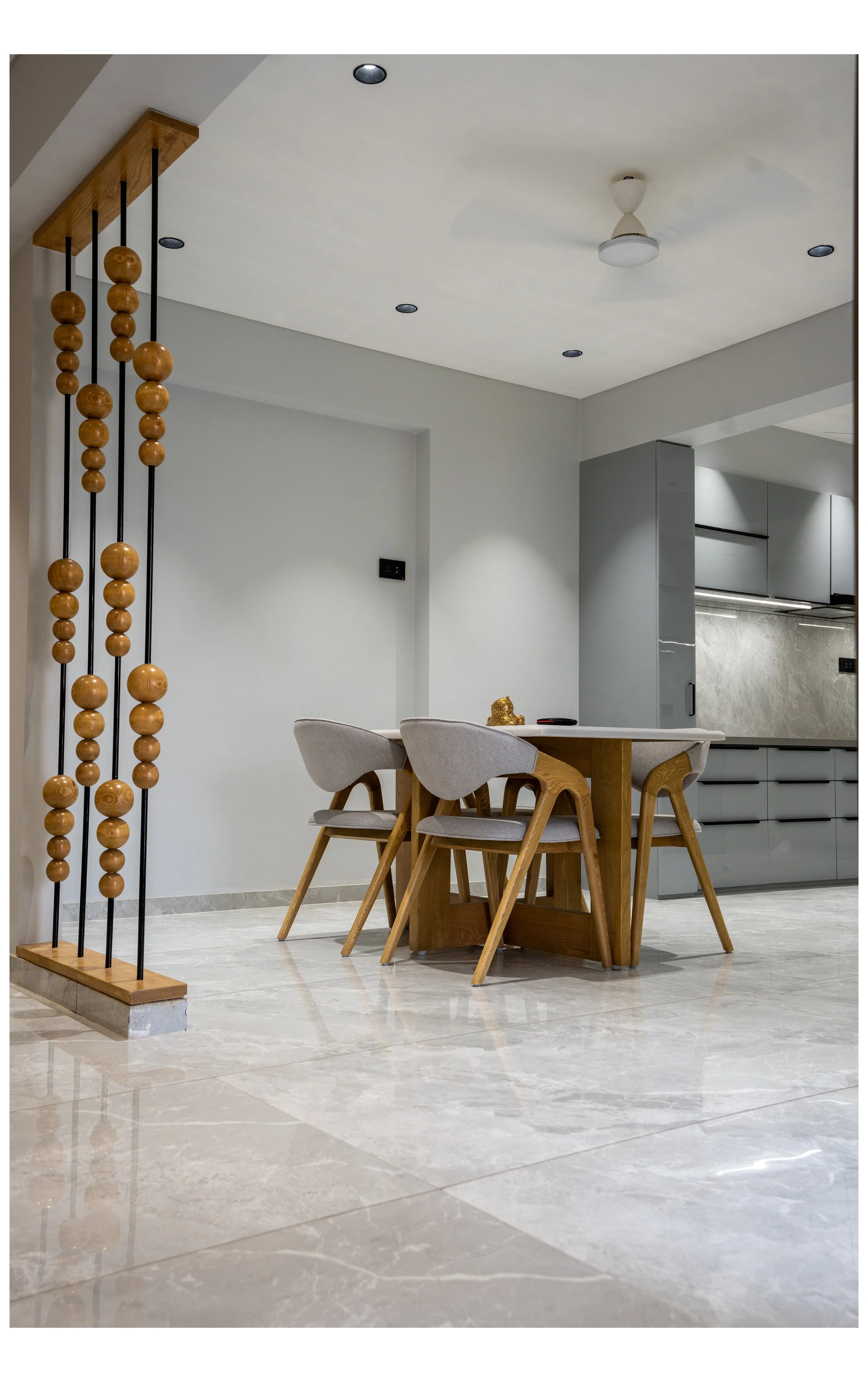 Modern dining area with a wooden table surrounded by four gray cushioned chairs. To the left, a decorative room divider with vertical black rods and wooden spheres. The floor is polished marble, and the kitchen with gray cabinets is visible in the ba