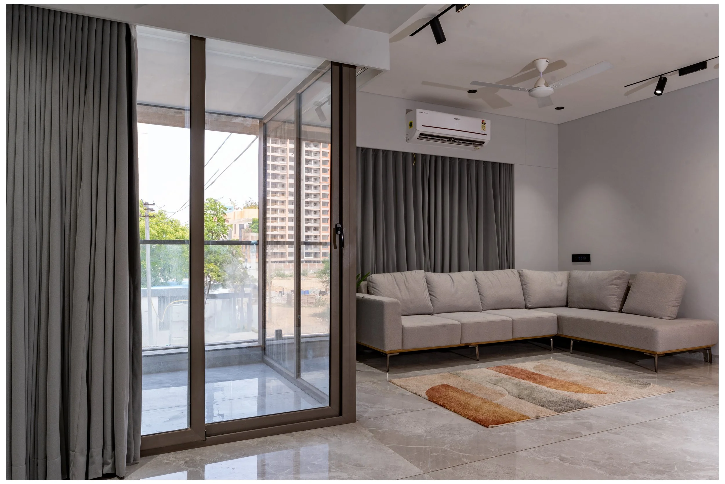 Modern living room with a beige sectional sofa, glass sliding door with gray curtains, small area rug, air conditioner, ceiling fan, and track lighting, overlooking a balcony with city view.