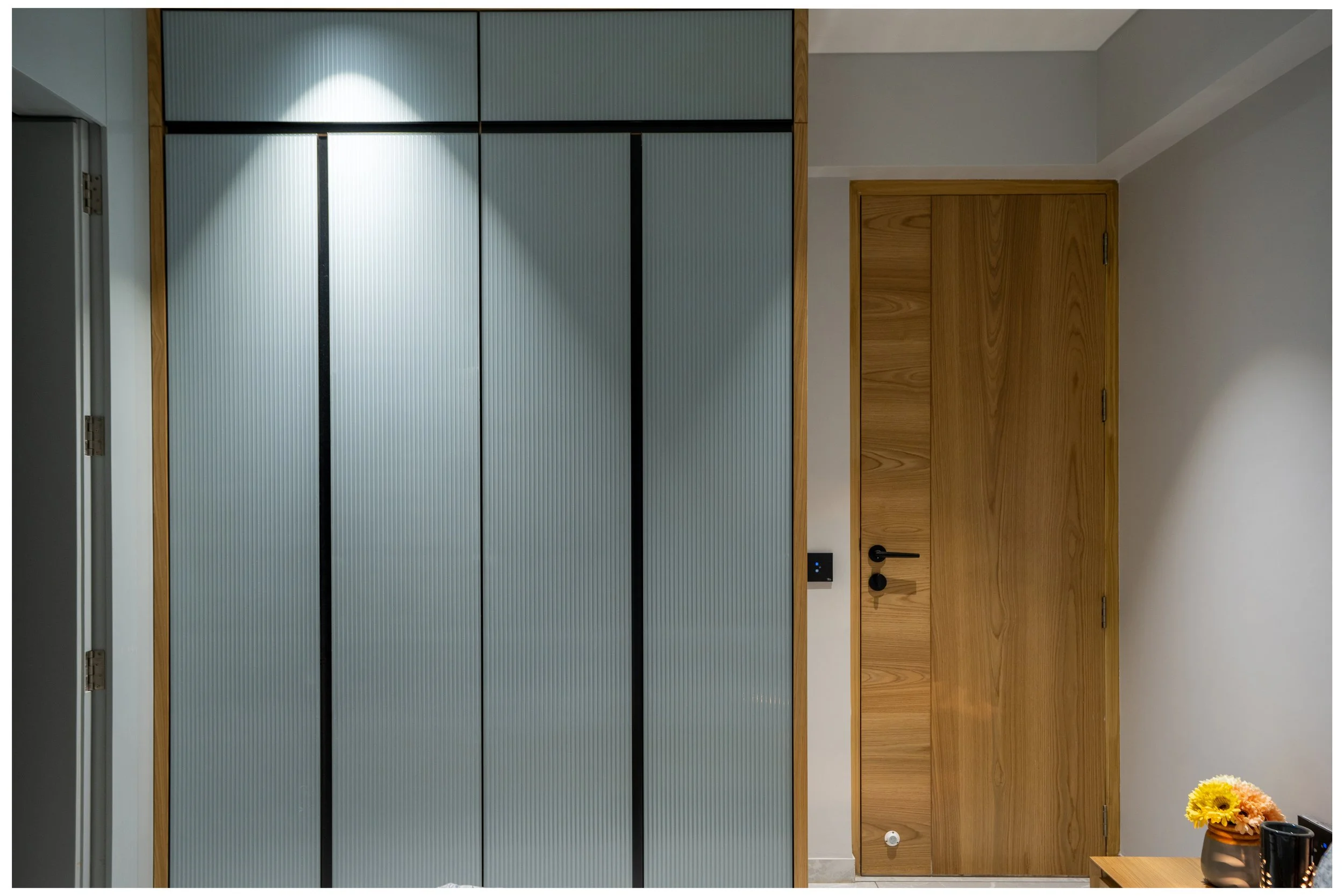 Interior of a modern room with a textured gray sliding door closet on the left, and a wooden door on the right. A small table with a vase of yellow and peach flowers is partially visible in the bottom right corner.