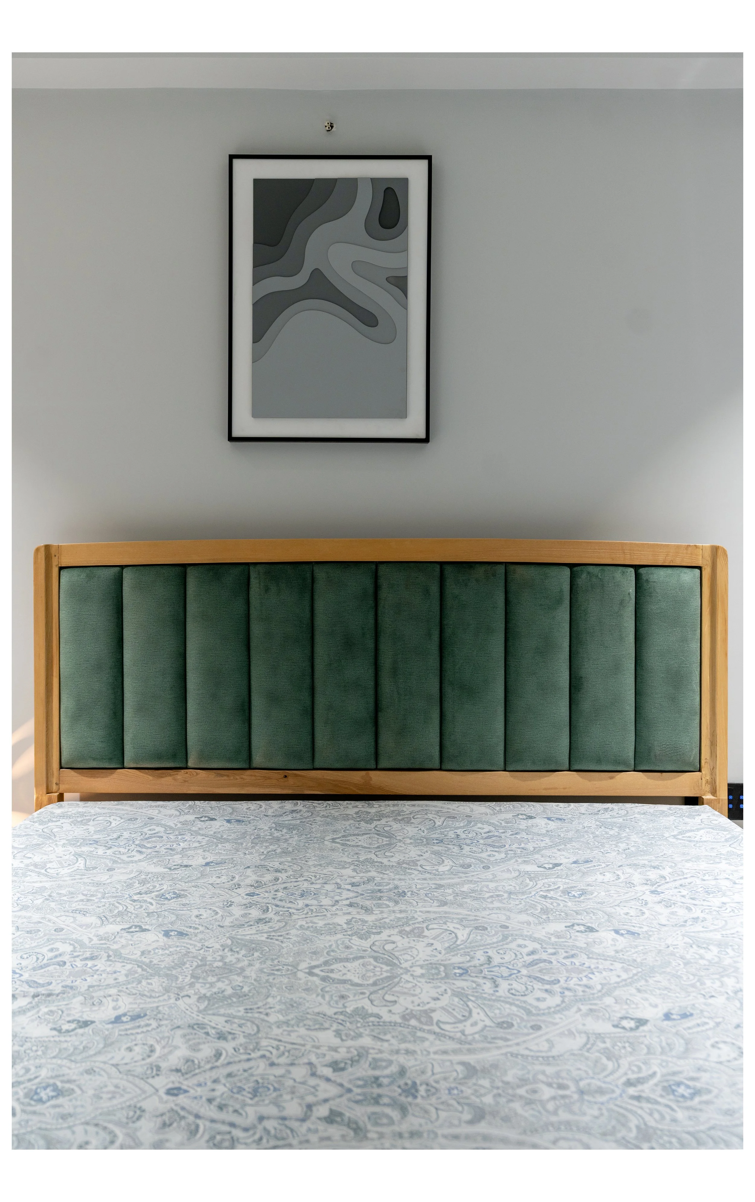 A bedroom featuring a bed with a light wood frame and green upholstered headboard, a quilted white and gray patterned bedspread, gray wall art with abstract shapes, and a light gray wall.