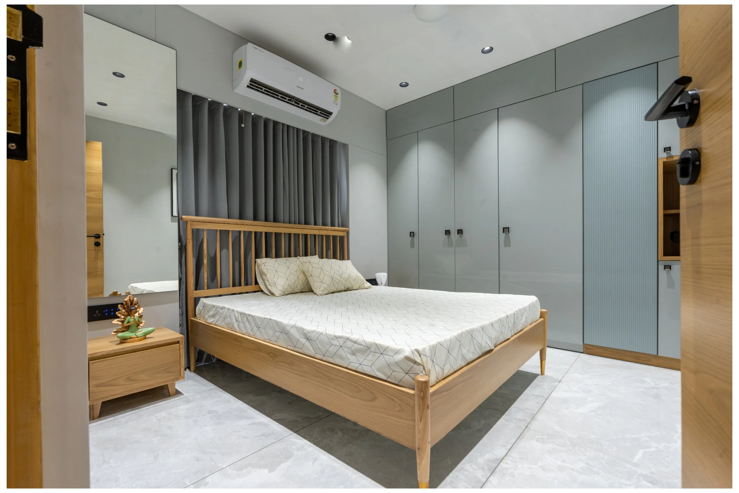 Modern bedroom with wooden bed frame, beige bedding, beige pillows, built-in gray wardrobe, wall mirror, small wooden bedside table with a decorative item, air conditioning unit, gray curtains, tiled floor.